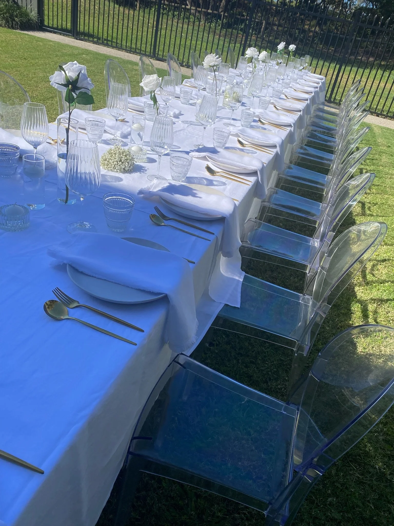 An outdoor dining table set with white tablecloth, white napkins, clear chairs, and glassware including wine glasses and water glasses, with some flowers as decoration.