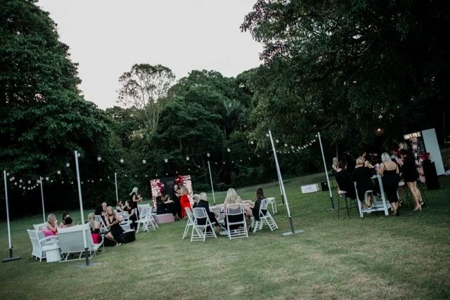 Guests sitting at tables and standing at an outdoor event in a grassy area with string lights and trees.