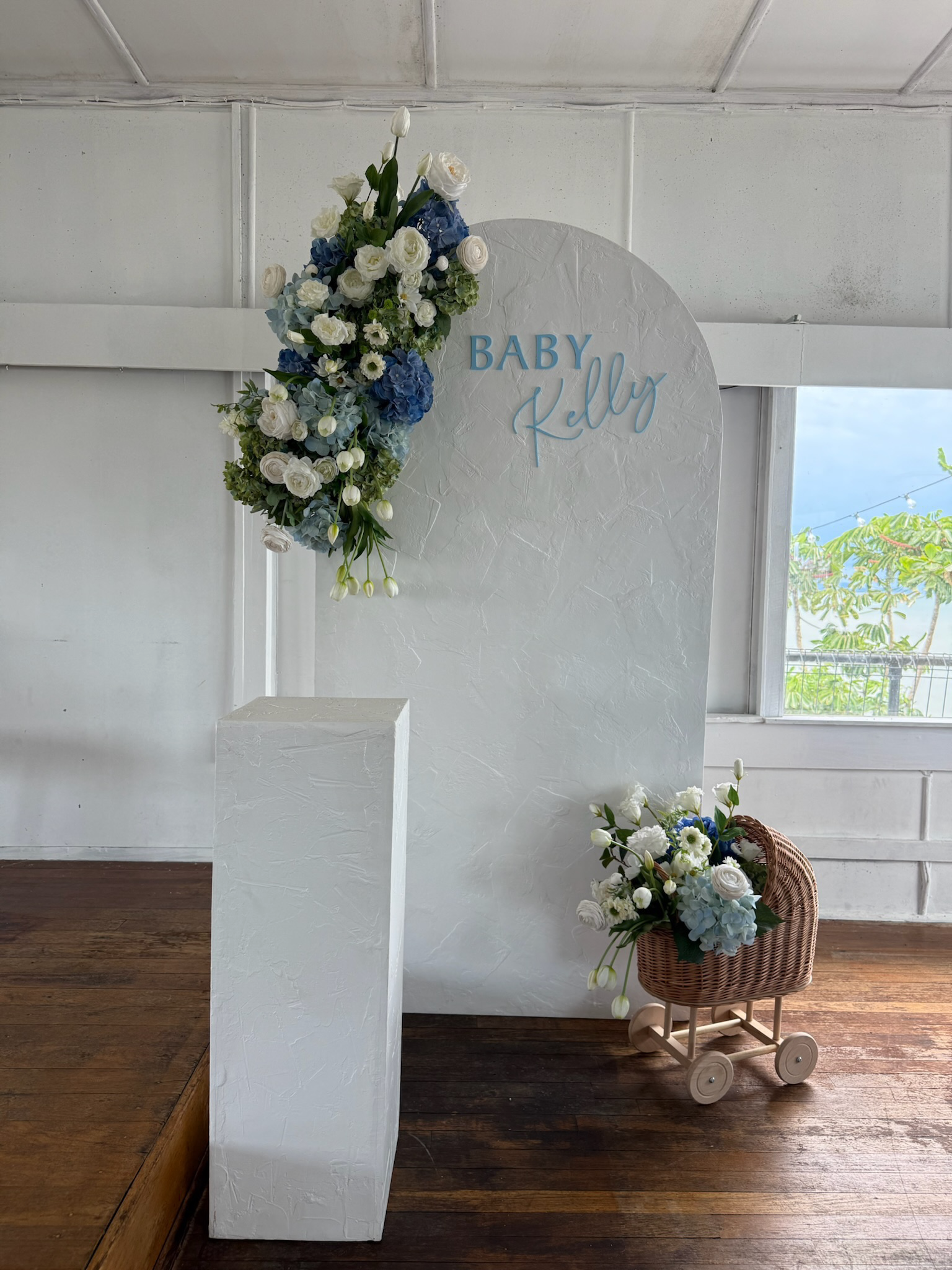 A white decorative backdrop with a blue sign that says 'Baby Kelly,' adorned with a floral arrangement of white and blue flowers, with a small cart filled with similar flowers next to it, set against a wall with a window showing outdoor greenery.