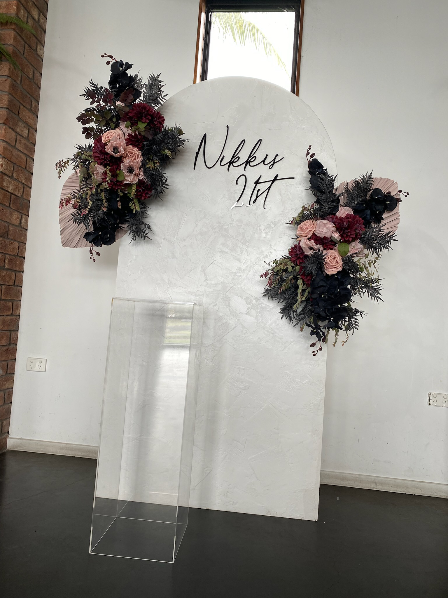 Decorative white backdrop with floral arrangements on either side, handwritten text reading "Nikkys 21st", and a clear podium in front. Free-standing window in the background.