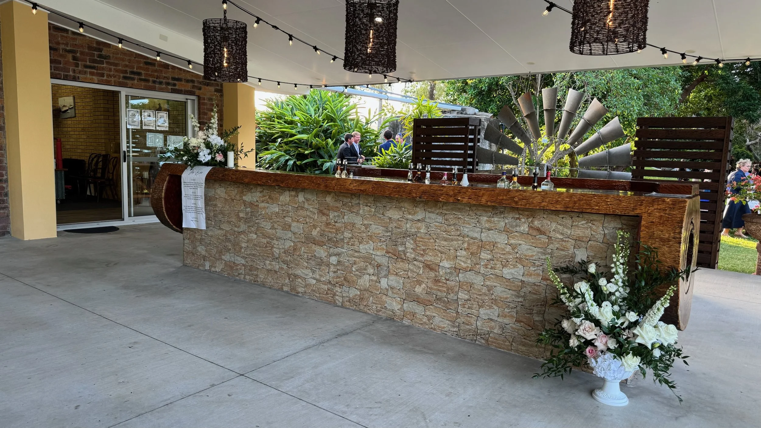 A decorated outdoor event space with a stone and wood bar, floral arrangements, string lights, and some people in suits.