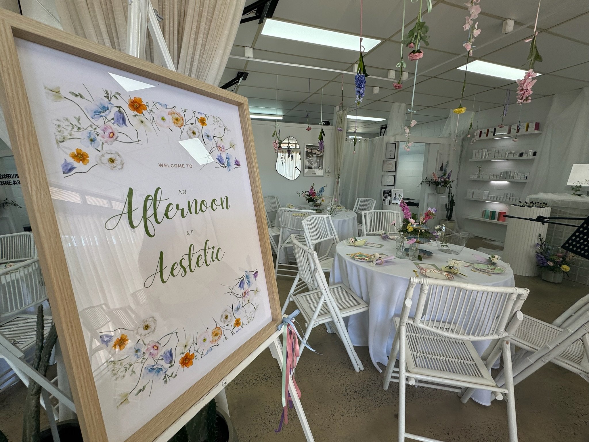 Decorated event space with round tables covered in white tablecloths, floral centerpieces, and white chairs. Hanging floral decorations from the ceiling and a sign welcoming guests to a-themed gathering.