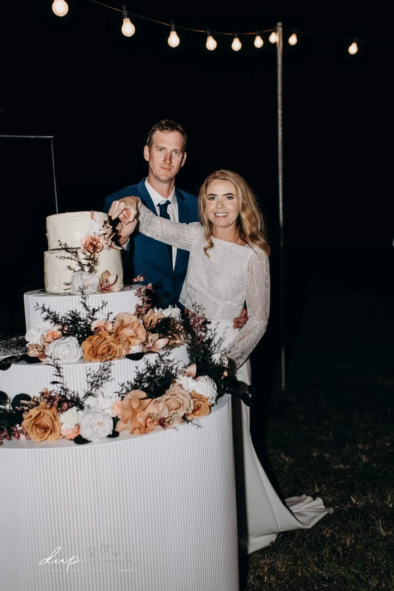 A bride and groom cut a wedding cake decorated with flowers and greenery at their wedding reception, illuminated by hanging string lights at night.
