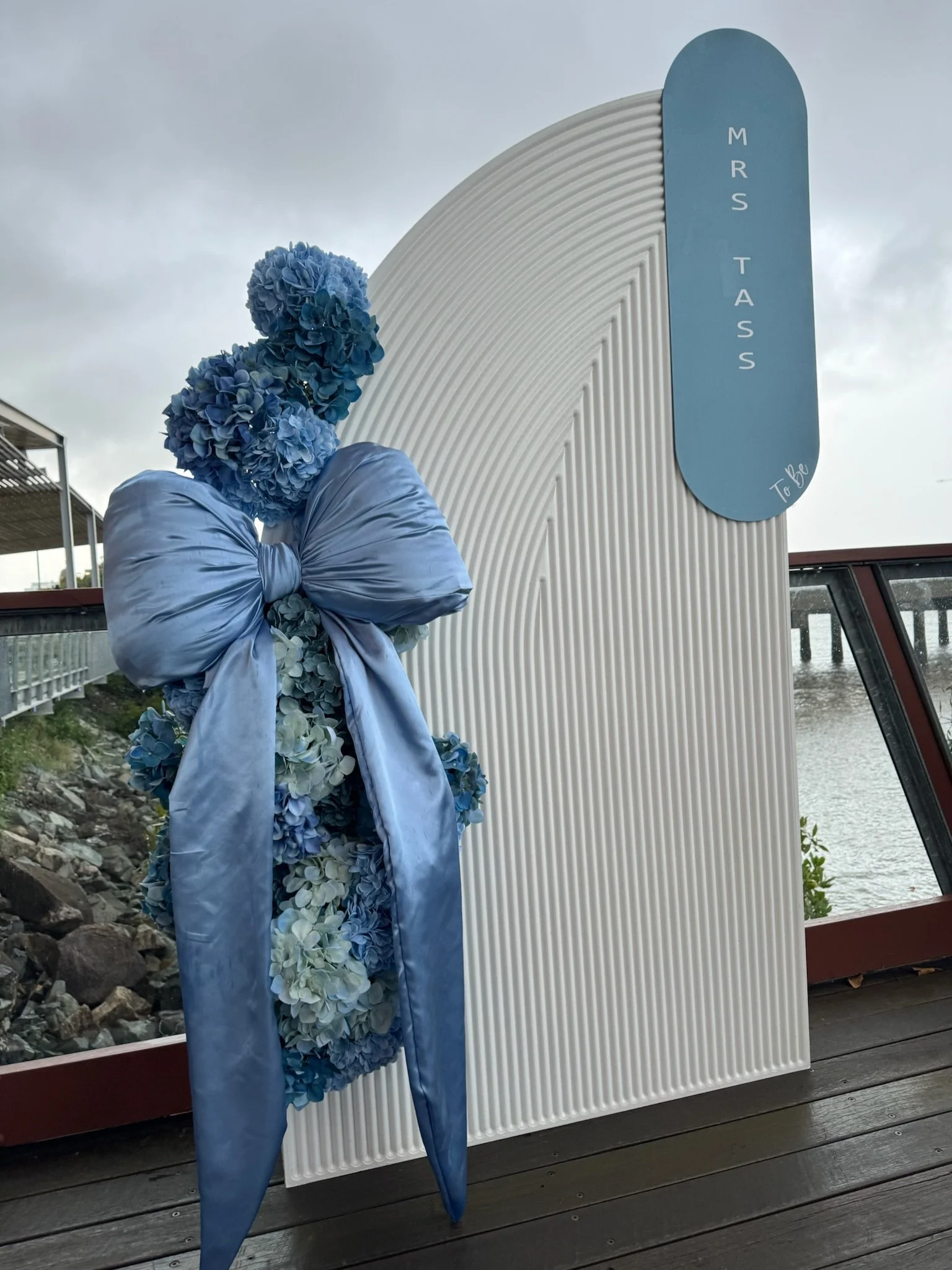 Blue floral tribute with hydrangeas and a satin bow, positioned next to an arch-shaped sign that reads 'MRS TASS' and 'To Be', near a water body and cloudy sky.
