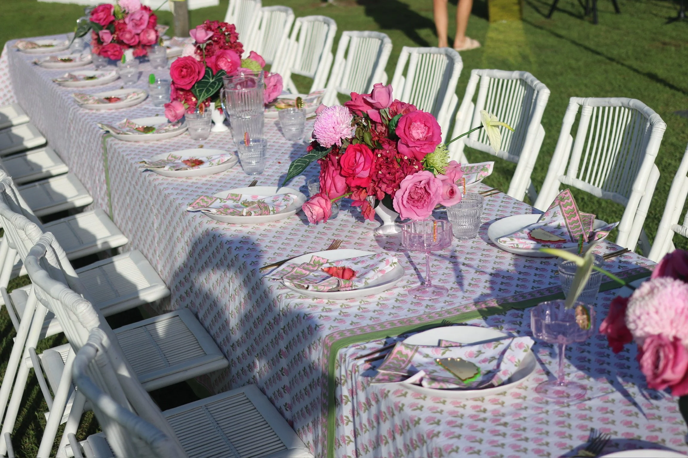 Long outdoor dining table set with pink floral arrangements, white chairs, and place settings on a grass lawn.