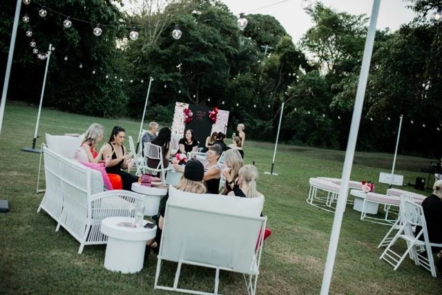 A group of women sitting together on white outdoor furniture at a garden party during the evening. Decorated with string lights and floral arrangements, with a black backdrop and trees in the background.