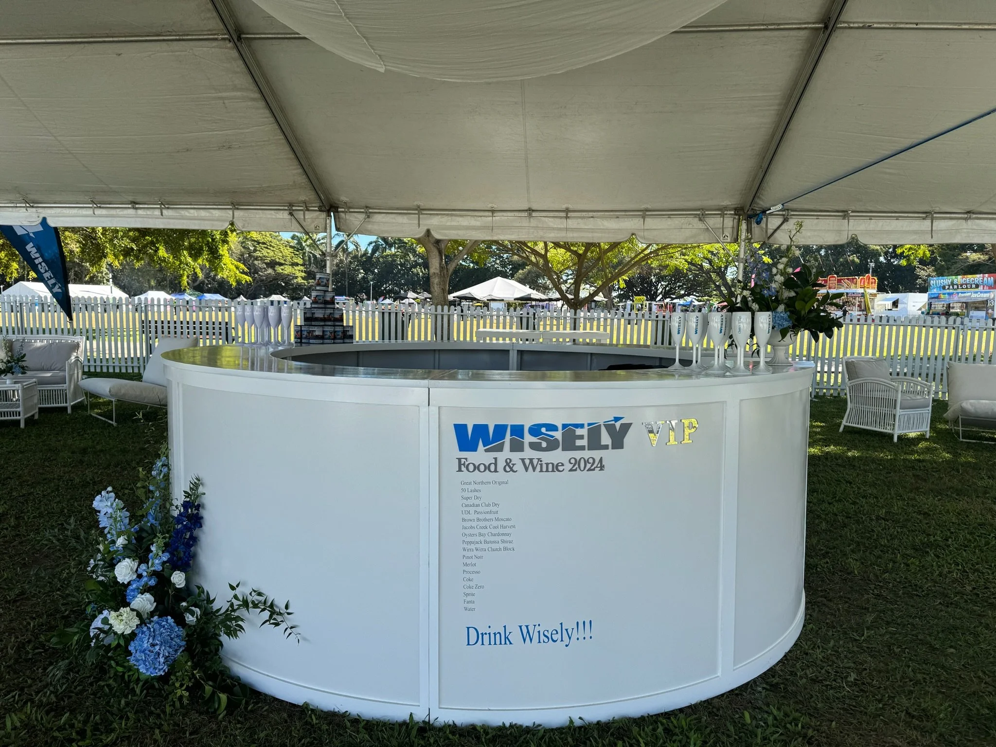 A white circular bar with Wisely Food & Wine 2024 signage, decorated with blue and white flowers, set up outdoors under a large canopy at a festival or fairground, with fields, trees, and tents in the background.