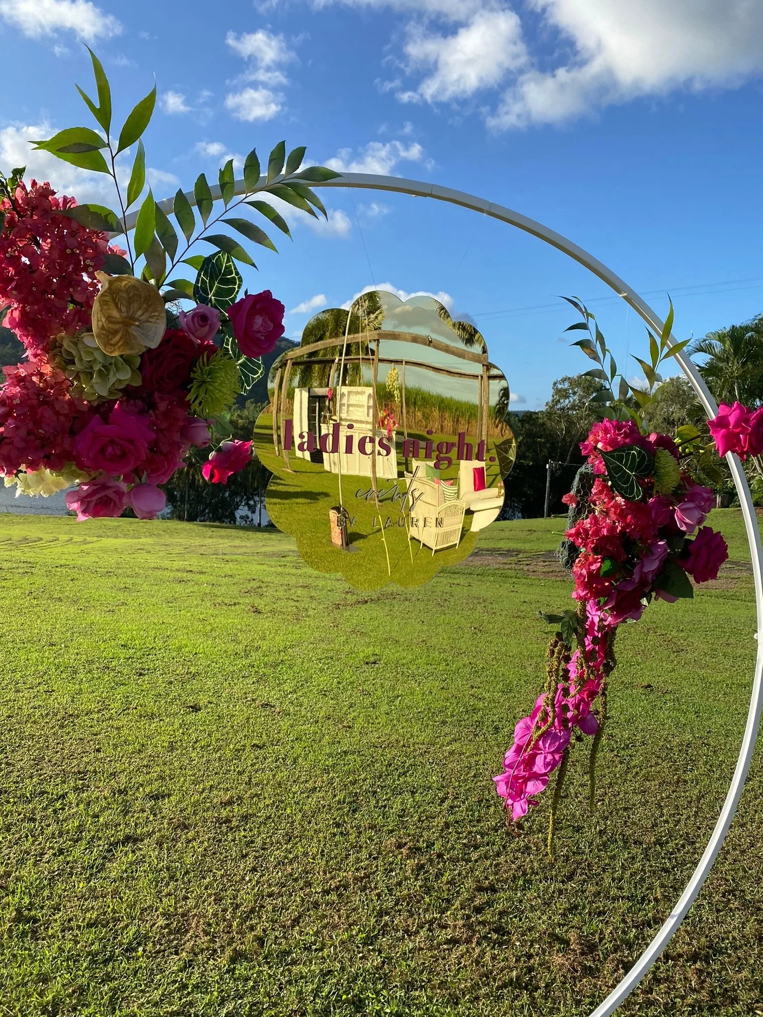 Decorative mirror with flowers and greenery in an outdoor setting, with a reflection of a house and a sign that reads "ladies night."