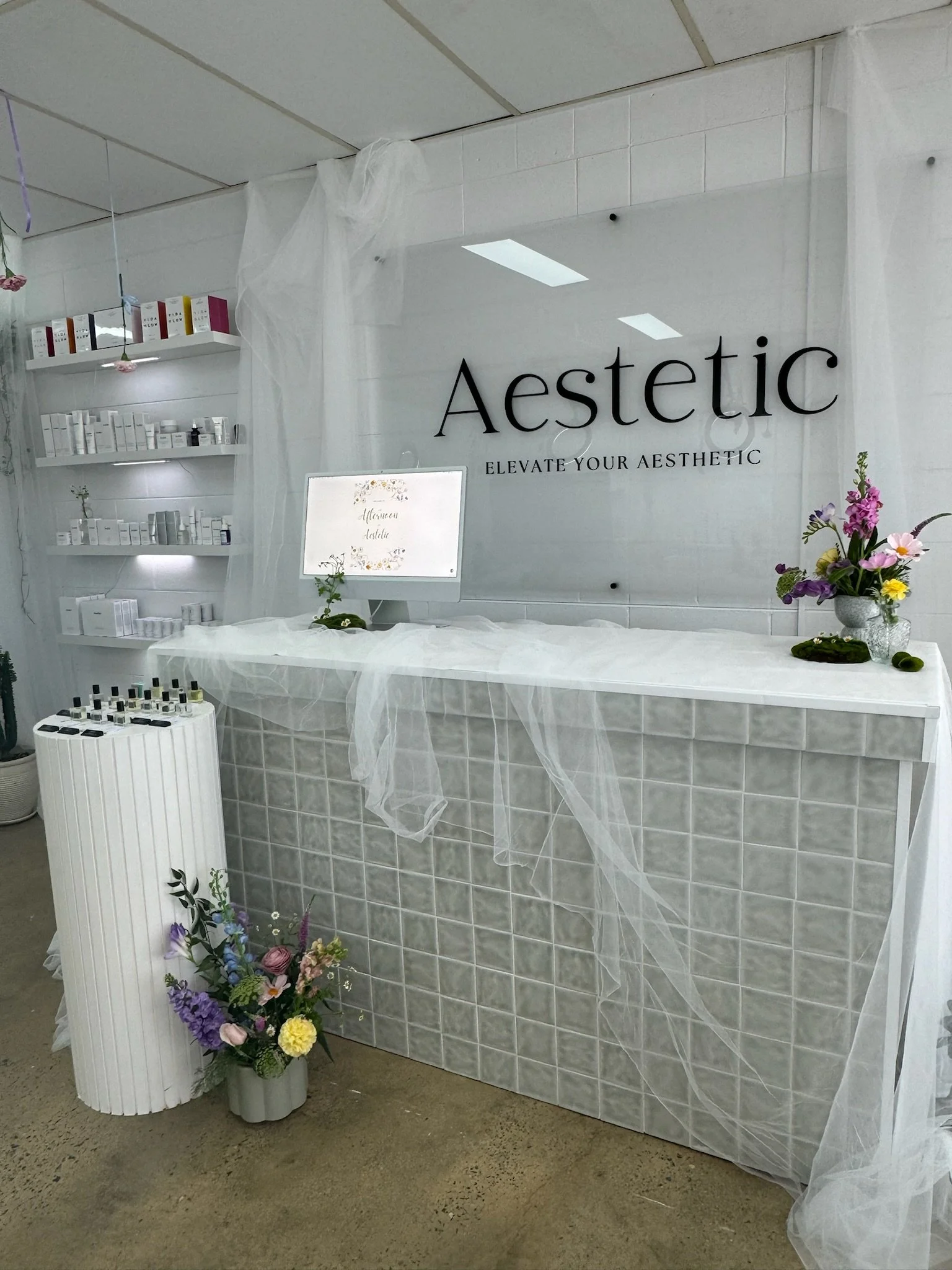 Minimalist aesthetic skincare or beauty salon reception area with white decor, a frosted glass sign reading 'Aesthetic, Elevate Your Aesthetic', colorful flower arrangements, and shelves with skincare products.