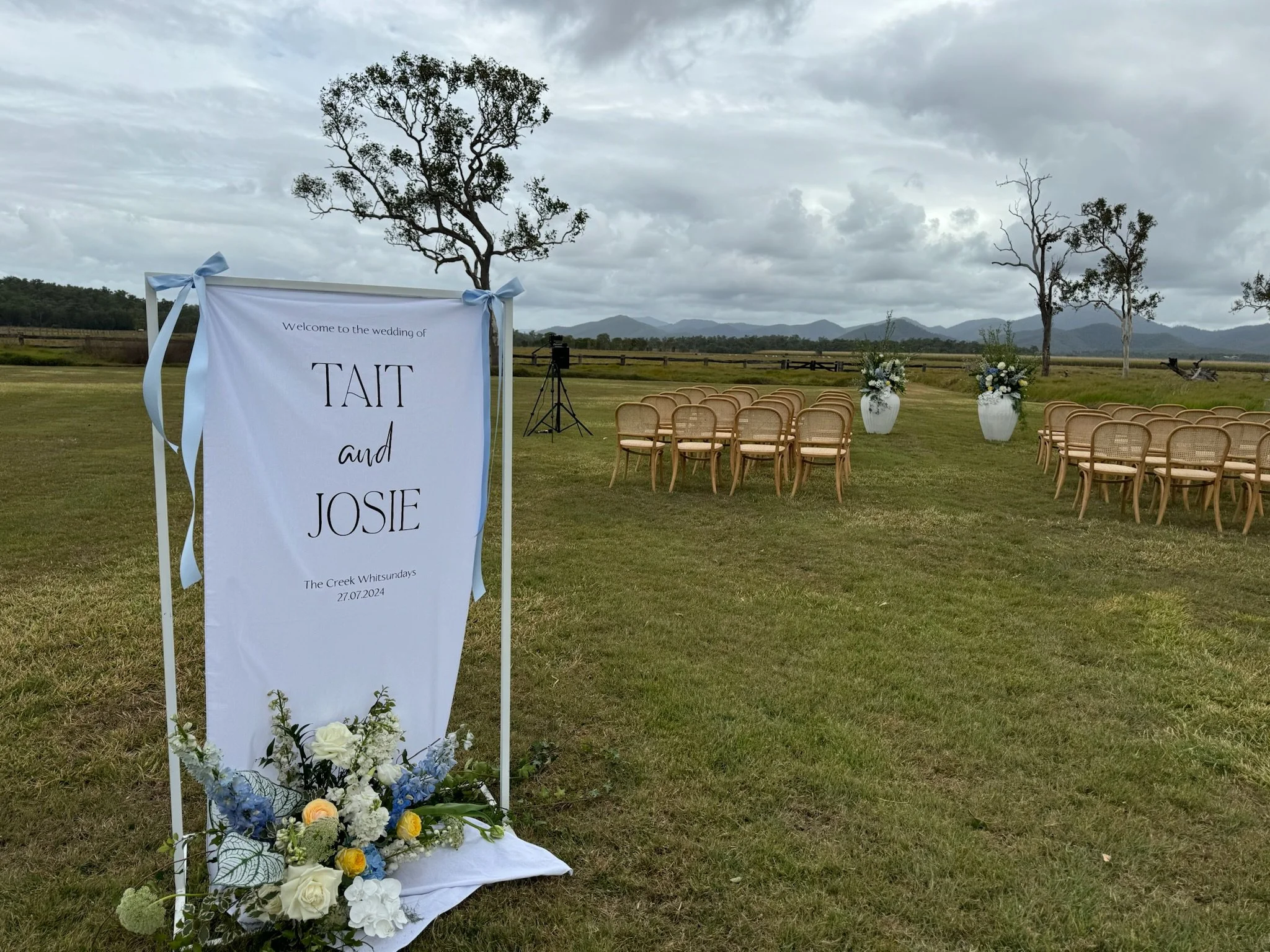 Outdoor wedding setup with rows of chairs facing a screen with a sign that reads, "Welcome to the wedding of Tait and Josie," decorated with white and blue flowers, under a cloudy sky with mountains in the background.