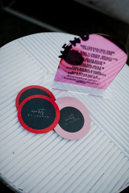 Three round coasters, two black with red borders and one pink with black text, placed on a white, ridged surface. A pink card with black text is positioned upright behind the coasters.