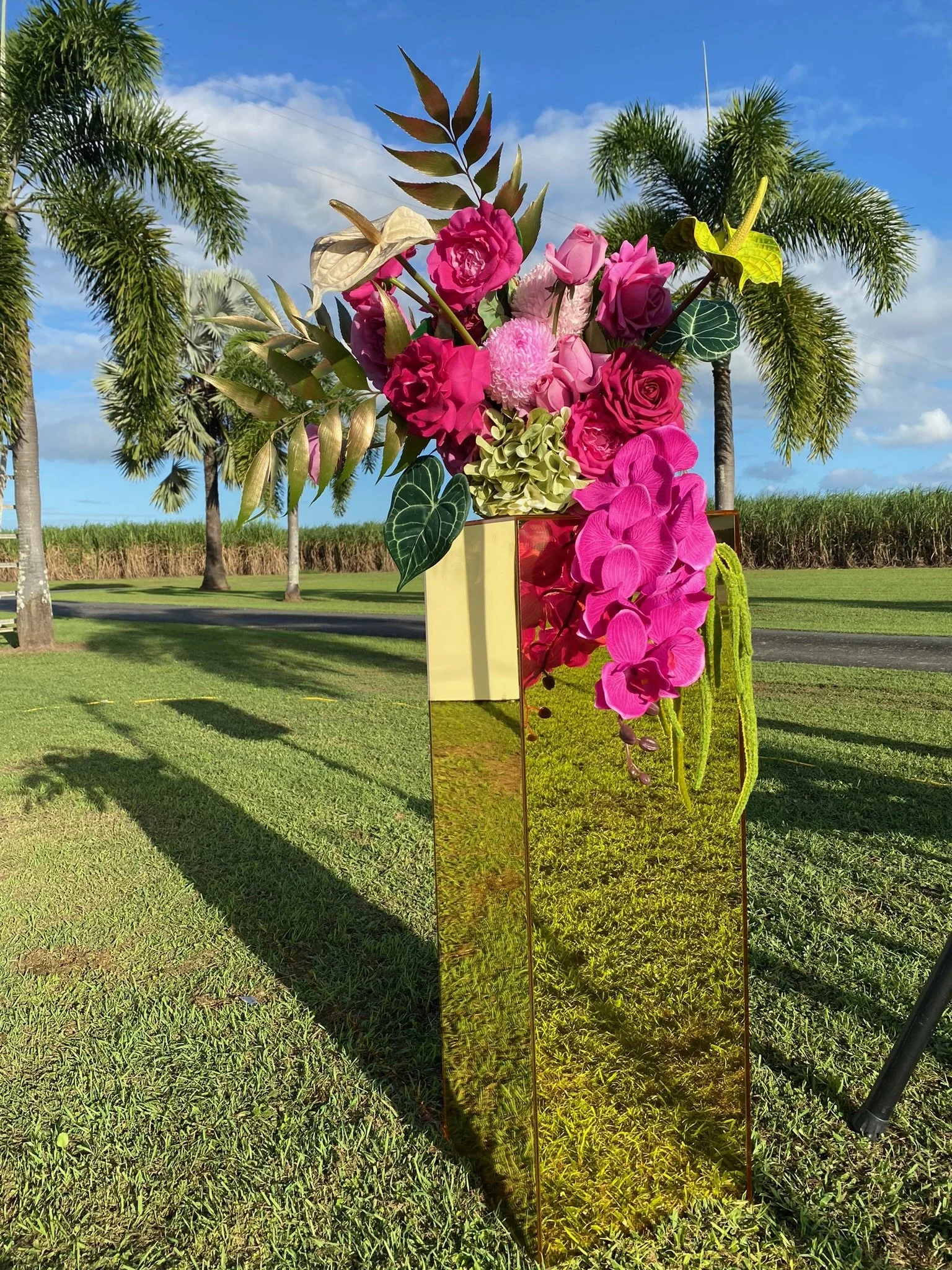 Colorful floral arrangement of pink and purple flowers in a reflective gold and bronze geometric vase outdoors on green grass with palm trees and blue sky in the background.