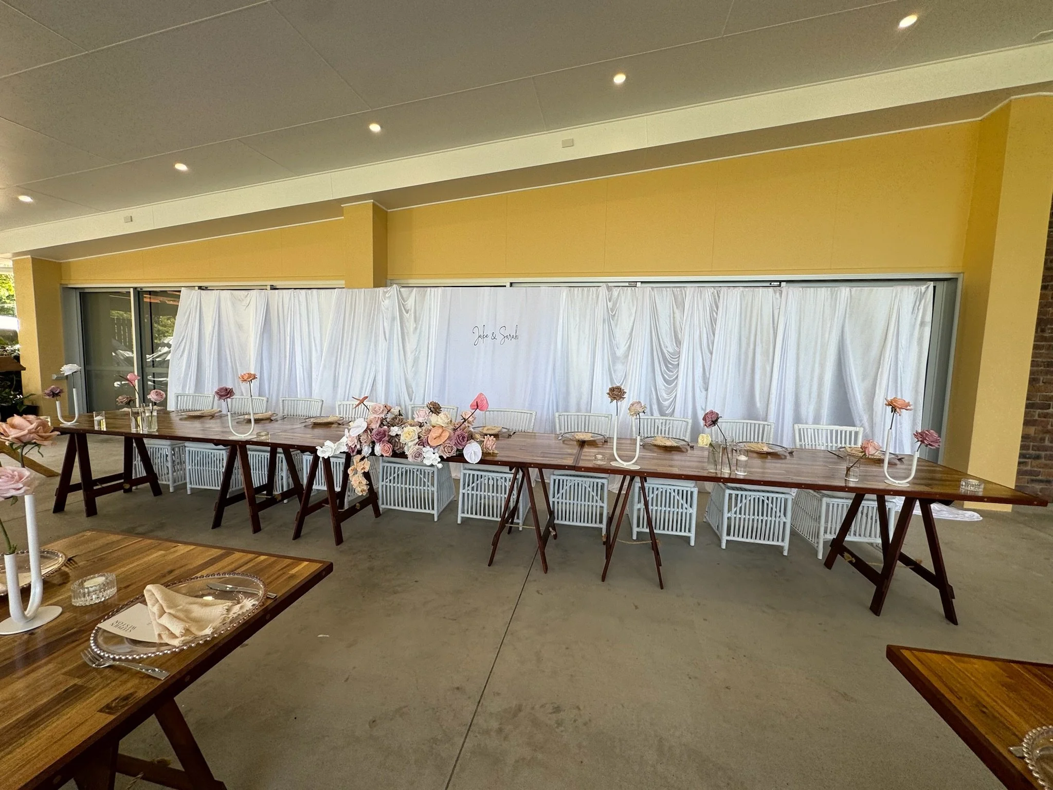 Wedding reception setup with a long wooden table decorated with pink and white flowers in vases, white chairs, and place settings on a concrete floor against a white draped backdrop with the names 'Jade & Sarah' written on it.