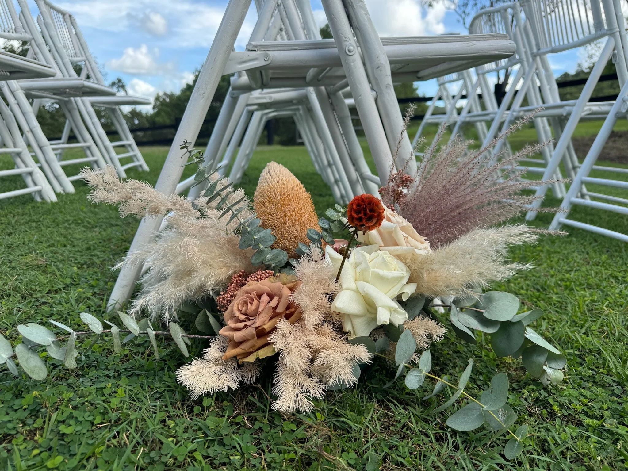 A floral arrangement with dried flowers and green leaves on grass, with white folding chairs stacked upside down and a partly cloudy sky in the background.