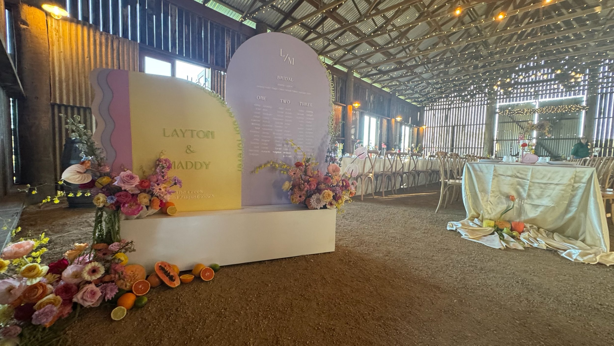 Wedding reception setup inside a rustic barn with floral arrangements, a decorative backdrop featuring names 'Layton & Maddy', chairs, and tables arranged for guests, with warm lighting and natural light coming through the windows.