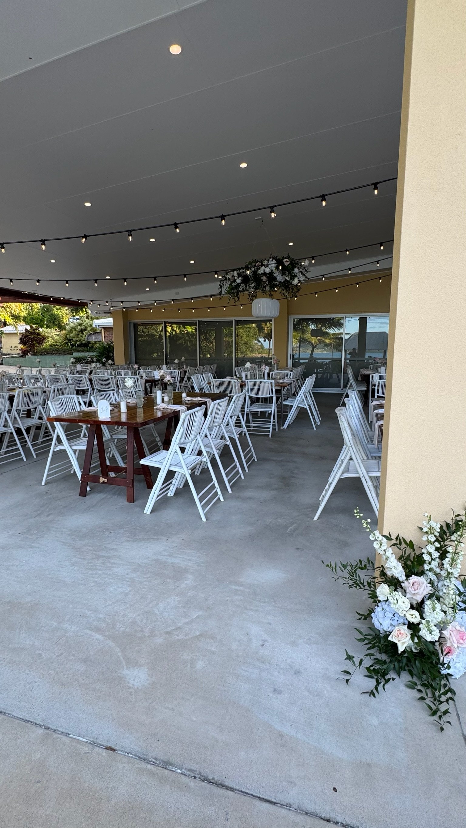 An outdoor event space decorated with string lights, a floral centerpiece hanging from the ceiling, and a flower arrangement on the ground near the corner, with tables and white chairs arranged for a celebration.