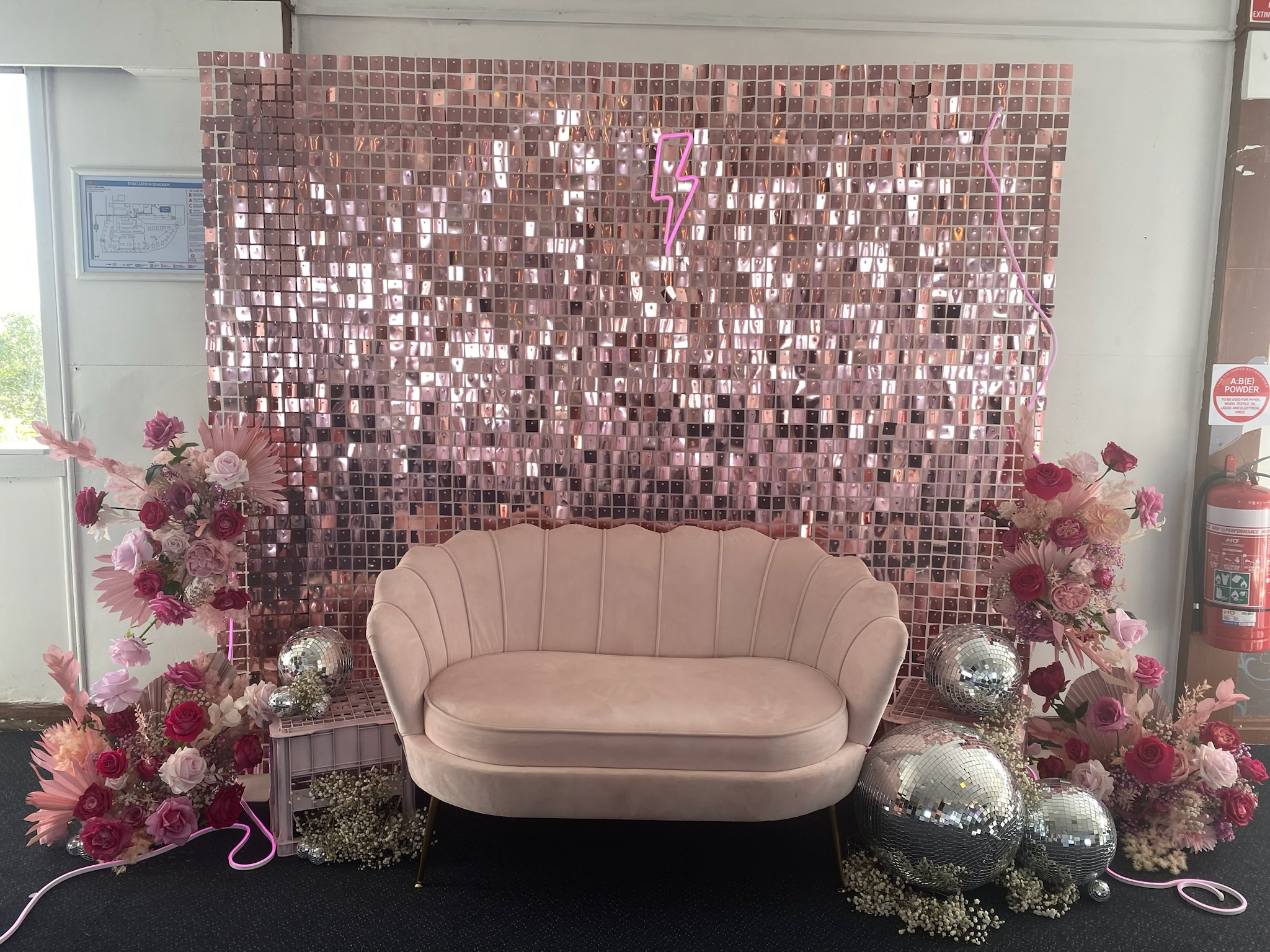 A pink velvet sofa situated in front of a pink mosaic wall with a neon pink lightning bolt light. Two floral arrangements with pink and red roses, pink dahlias, and baby's breath are on each side, with disco balls of various sizes on the floor beneat
