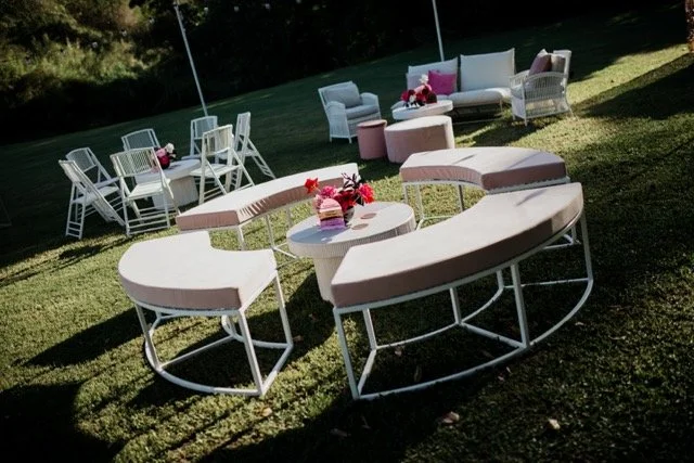 Outdoor seating arrangement with curved white benches and pink cushions, surrounded by flower bouquets on tables, set on a lawn with chairs and sofas in the background.