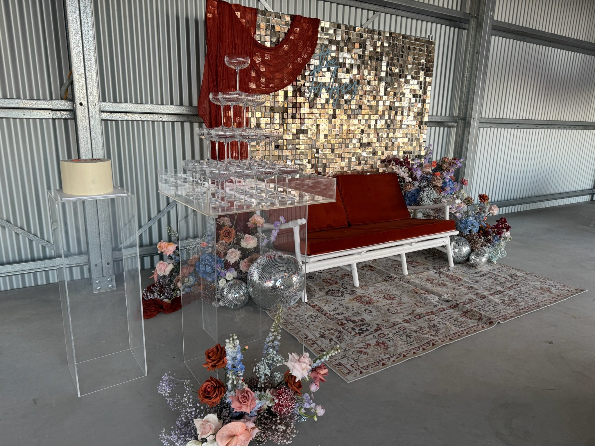 Decorative setup with a vintage red Cushioned loveseat, a flower arrangement, a backdrop of shiny mosaic tiles with cursive writing, a glass table with empty champagne glasses stacked to form a tower, silver disco balls, floral decorations, and a cre