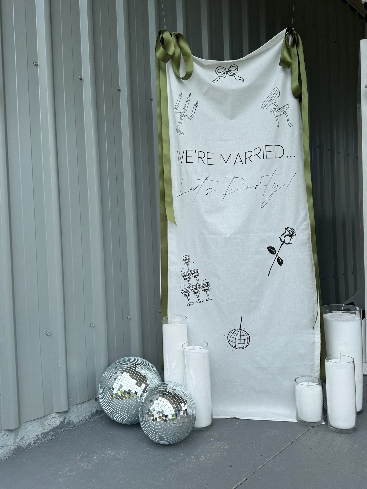 Wedding celebration decor with a white fabric banner that reads 'WE'RE MARRIED... Let's Party!' surrounded by candles, disco balls, and illustrations of a candelabra, champagne glasses, a rose, and a disco ball.