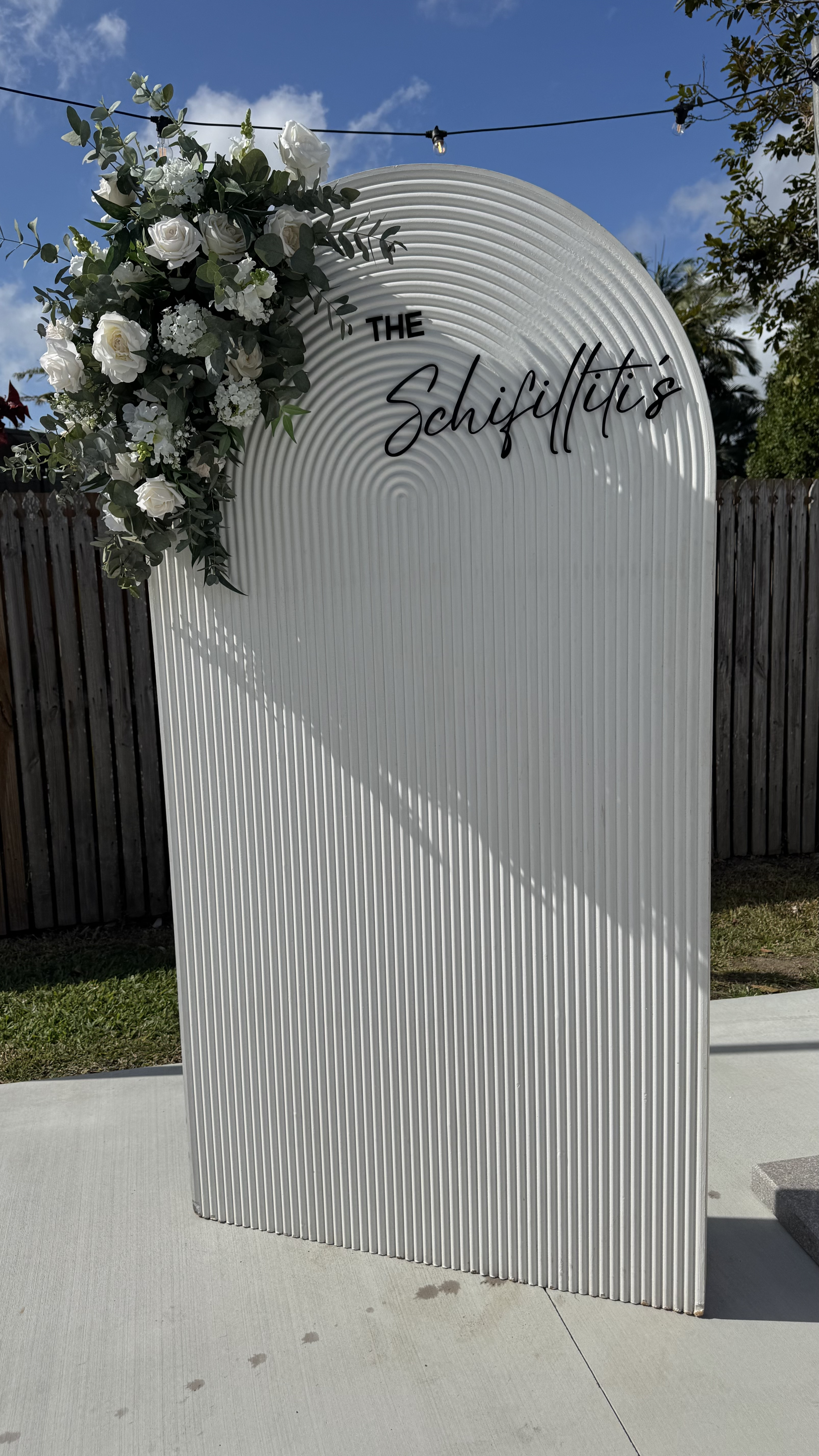 White standing board with floral decoration on the top left corner reading 'The Shifitis' in black cursive and capital letters, outdoors with a fence and trees in the background.