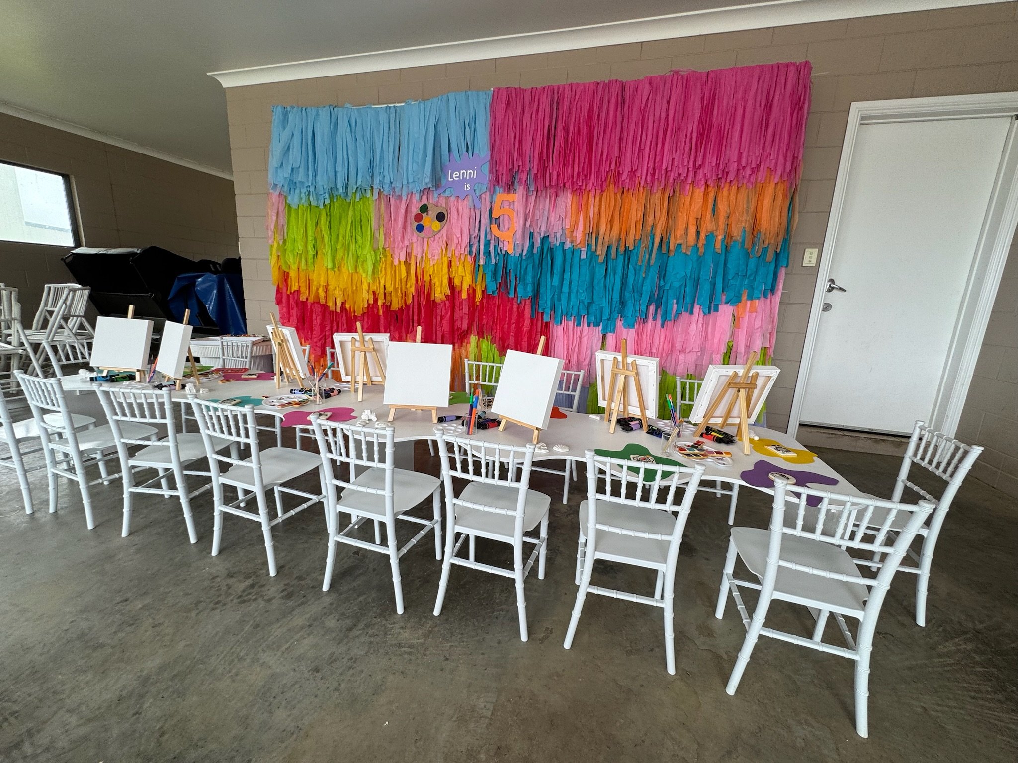 Party table with white chairs set up for a painting activity, decorated colorful tissue paper backdrop, signs with the name Lenni and a number 5, and art supplies on the table.
