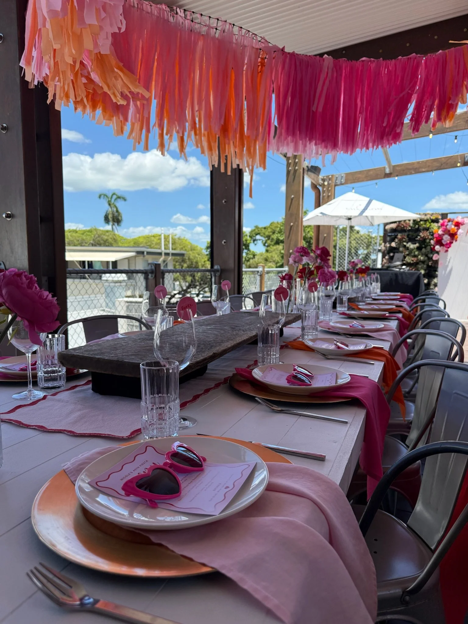 Decorated outdoor dining table with pink and orange tassel banners hanging above, pink flowers, place settings with pink sunglasses, and a sunny sky with clouds in the background.