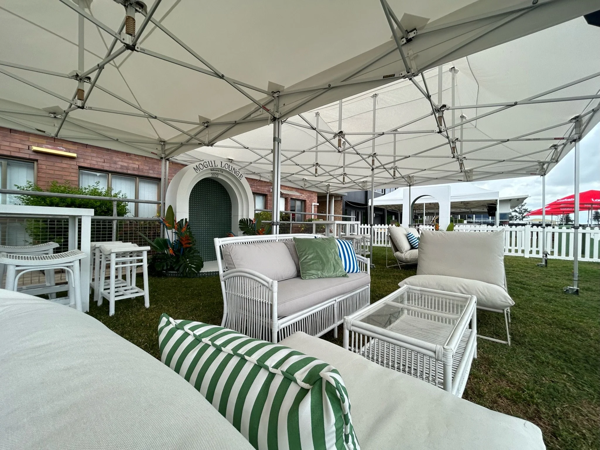 Outdoor lounge area under a canopy with white patio furniture, cushions, and small tables, with a brick building and white picket fence in the background.