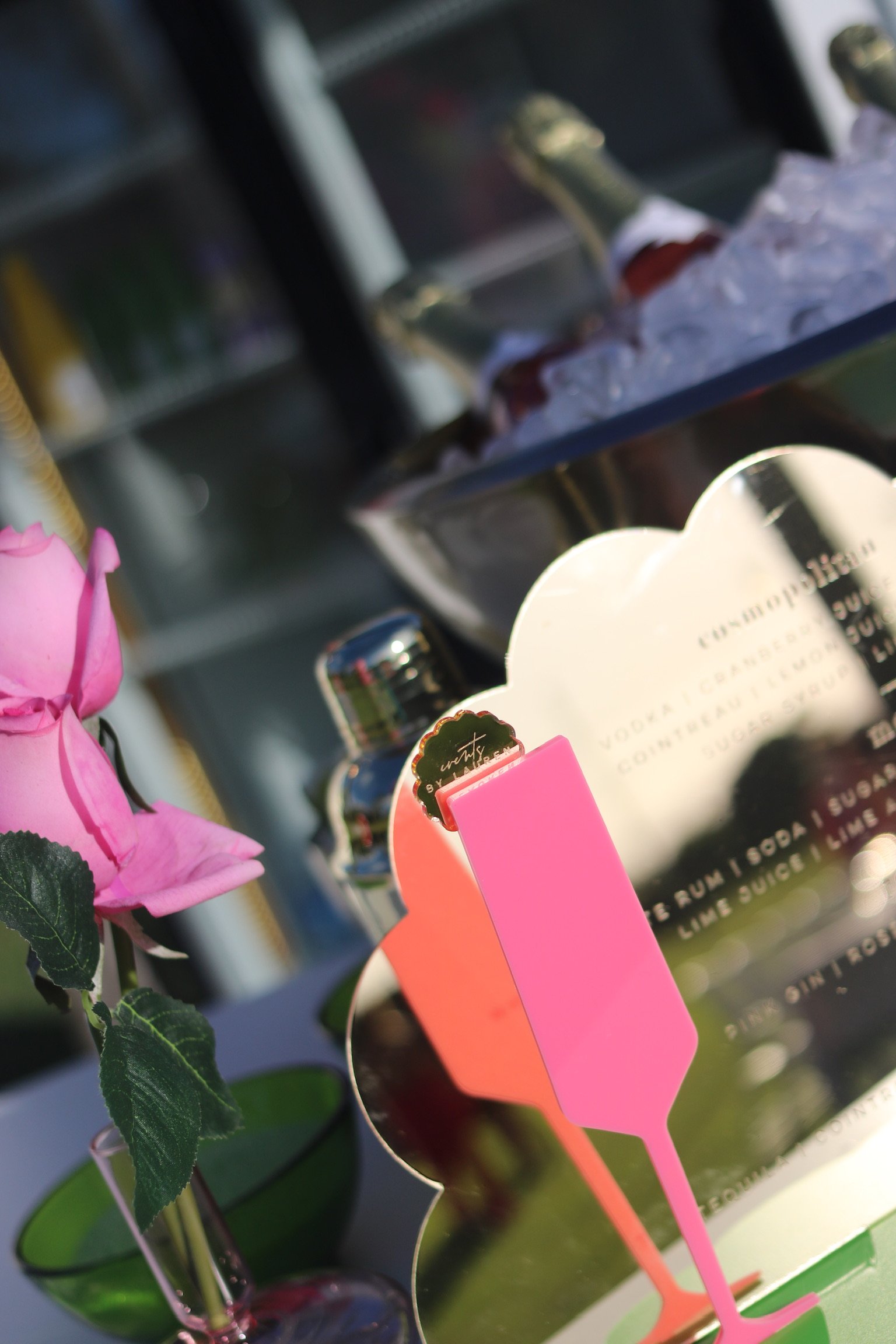 Close-up of a pink cocktail stirrer in front of a reflective card or sign with text, pink flower, and bottles in the background.