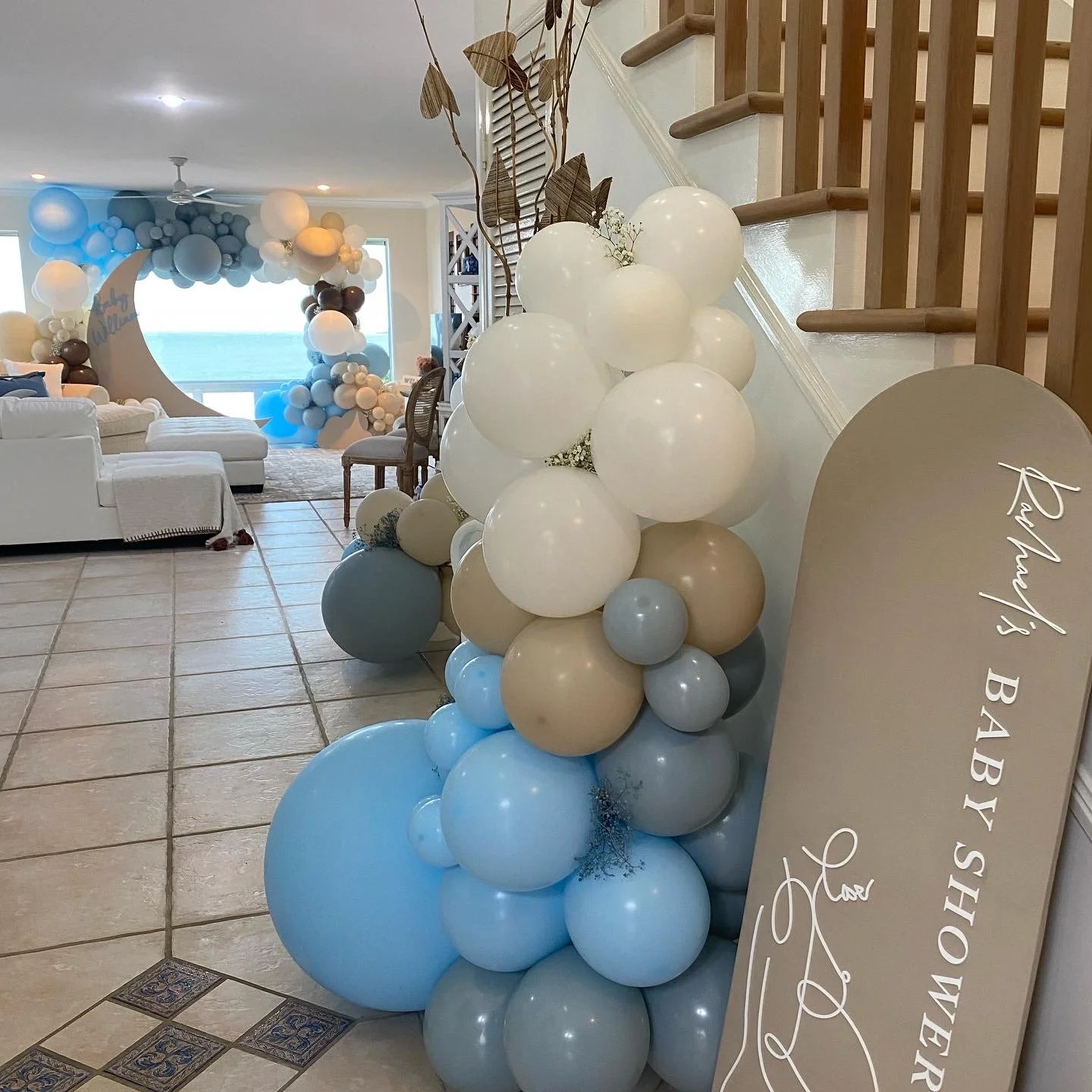Decorative balloon arrangement with white, beige, and blue balloons under a staircase and a sign that reads 'Rachel's Baby Shower' placed on the floor.