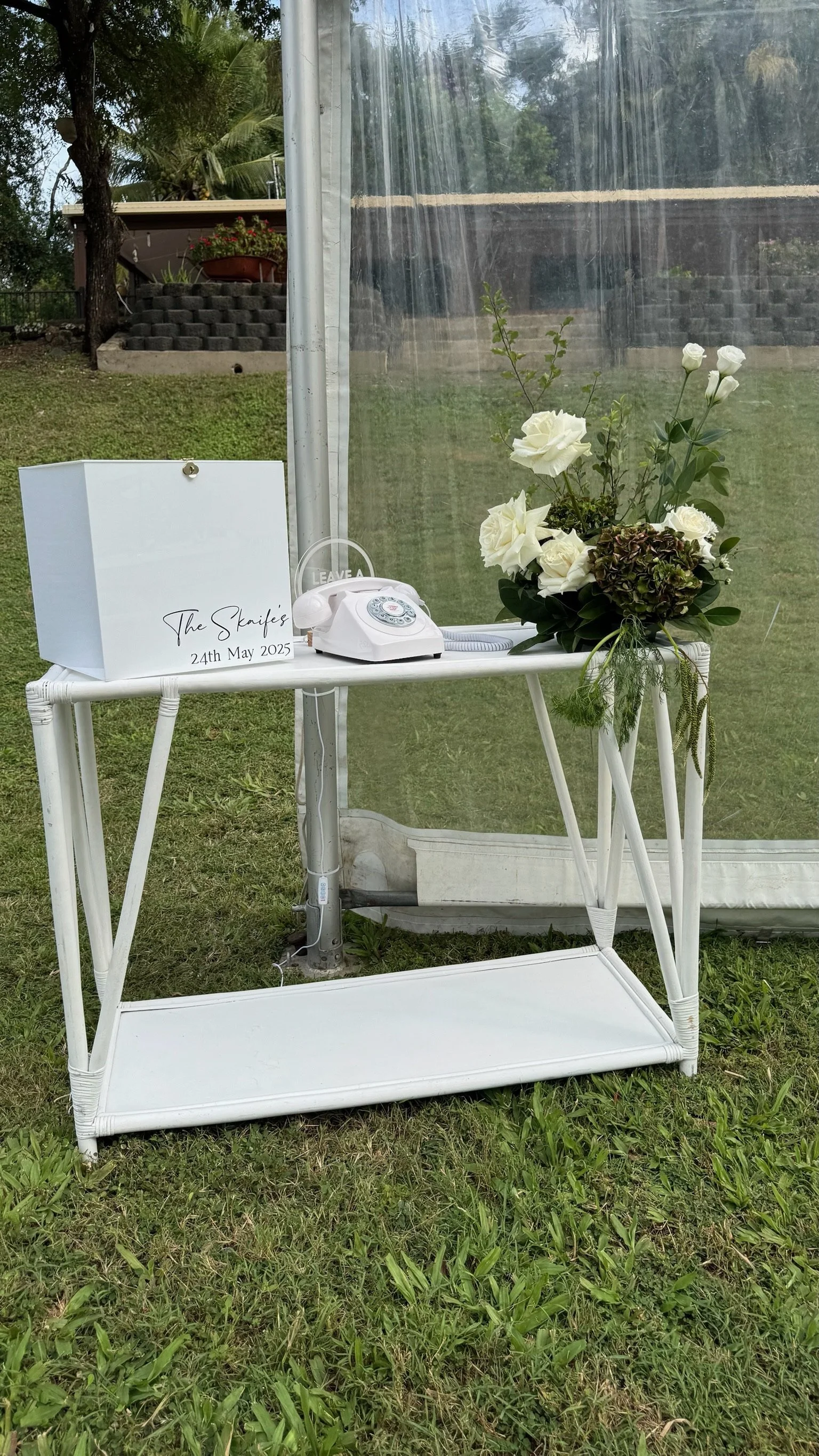 A white table with a vintage rotary phone, a white box labeled with a wedding date, and a floral arrangement with white roses and greenery outdoors on grass.