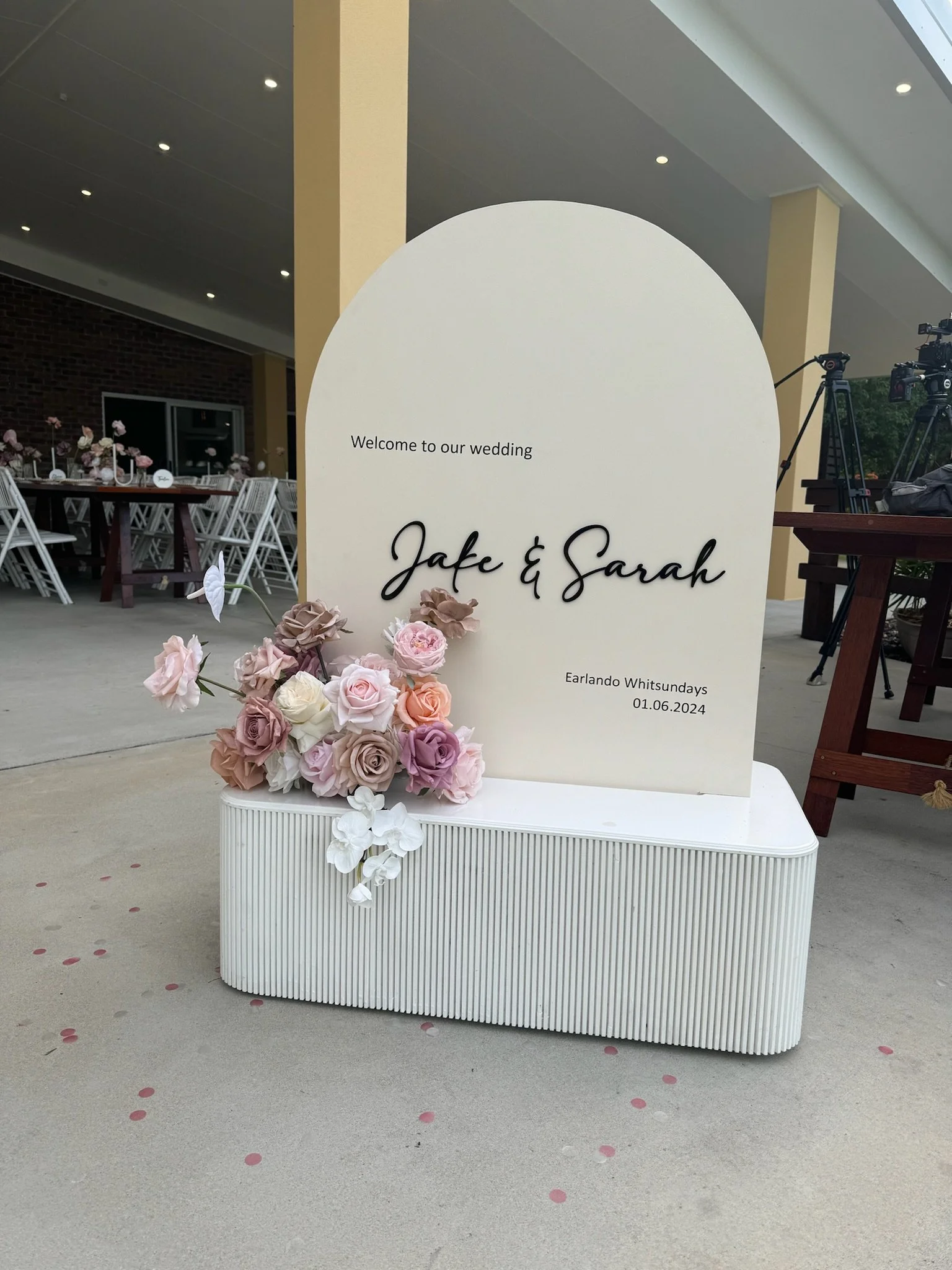 Wedding welcome sign with pink and white roses, off-white background, text welcoming guests to Jake and Sarah's wedding on June 1, 2024, placed outside near tables and cameras.