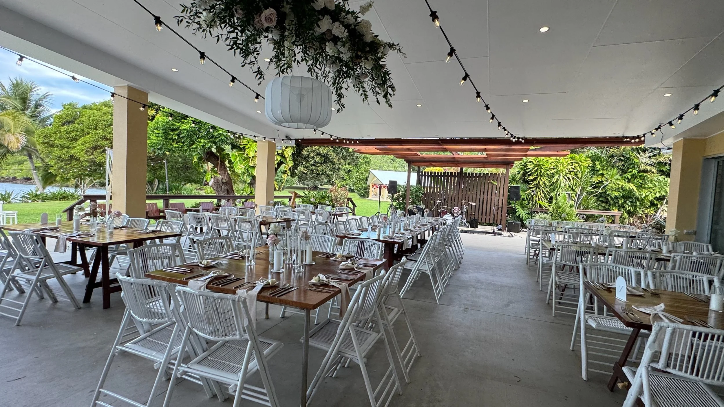 Setup for an outdoor event with long tables, white chairs, and table settings, under a covered patio with string lights and a view of greenery and water outside.