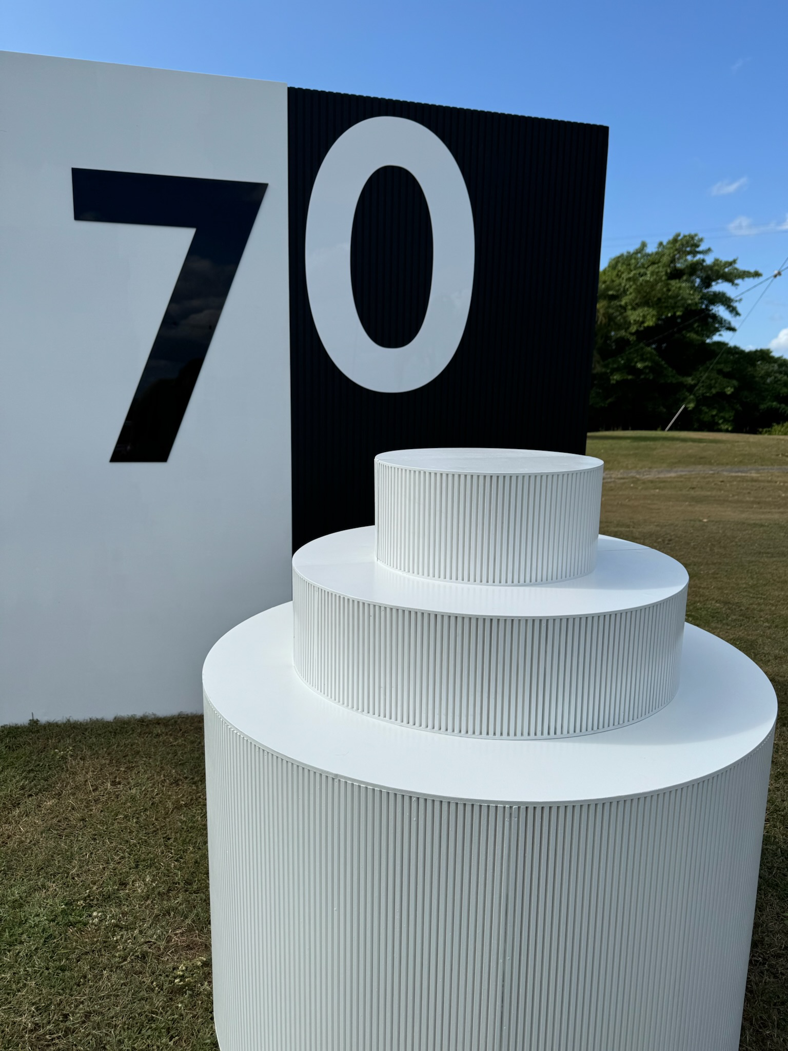 Black and white podiums with a large "70" sign behind them, set outdoors on a grassy field with trees and a blue sky in the background.