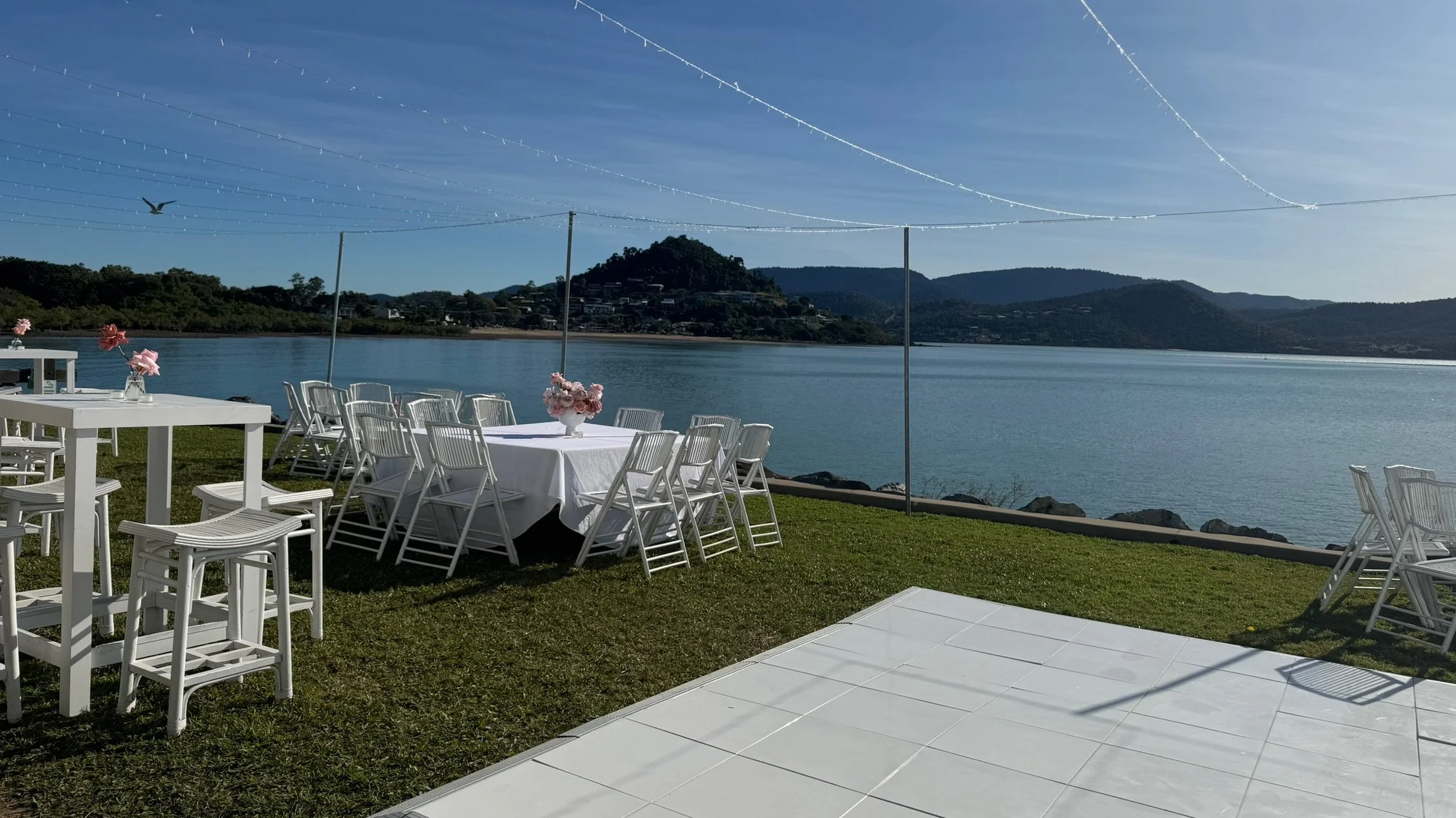Outdoor dining setup with white tables and chairs, decorated with pink flowers, by a lake with hills in the background under a clear blue sky.