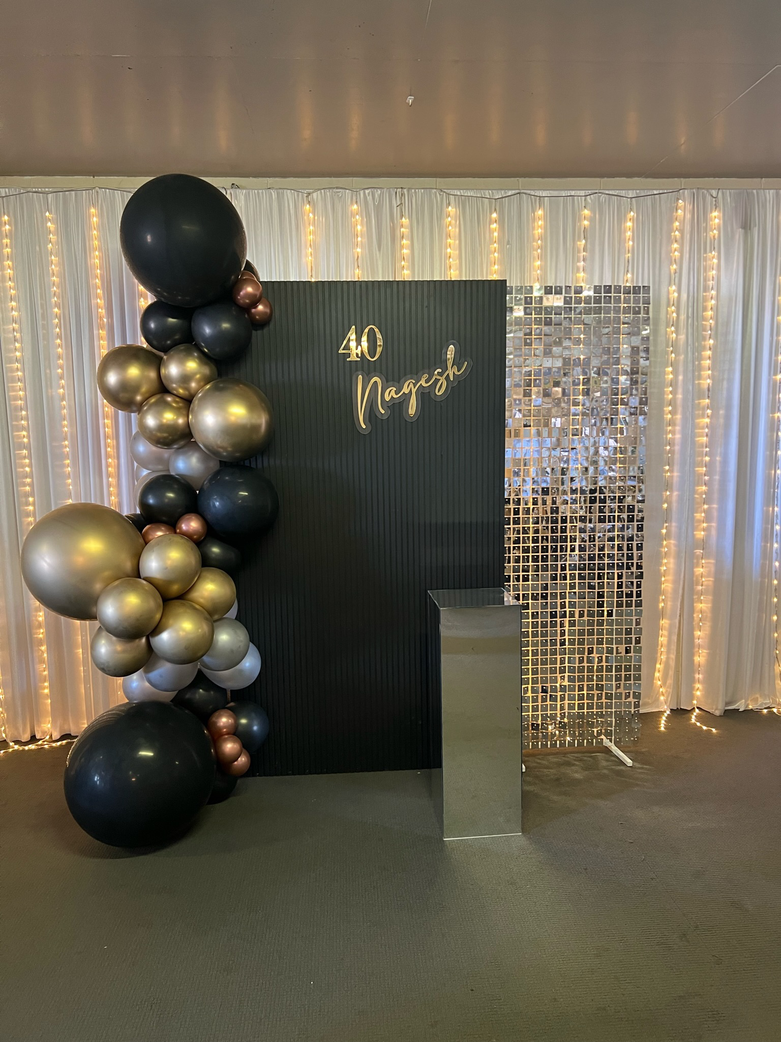 Decorative backdrop celebrating a 40th birthday, with black and gold balloons, a dark green panel with the text '40 Nagesh' in gold, white curtains with string lights, and a mirrored panel to the right.