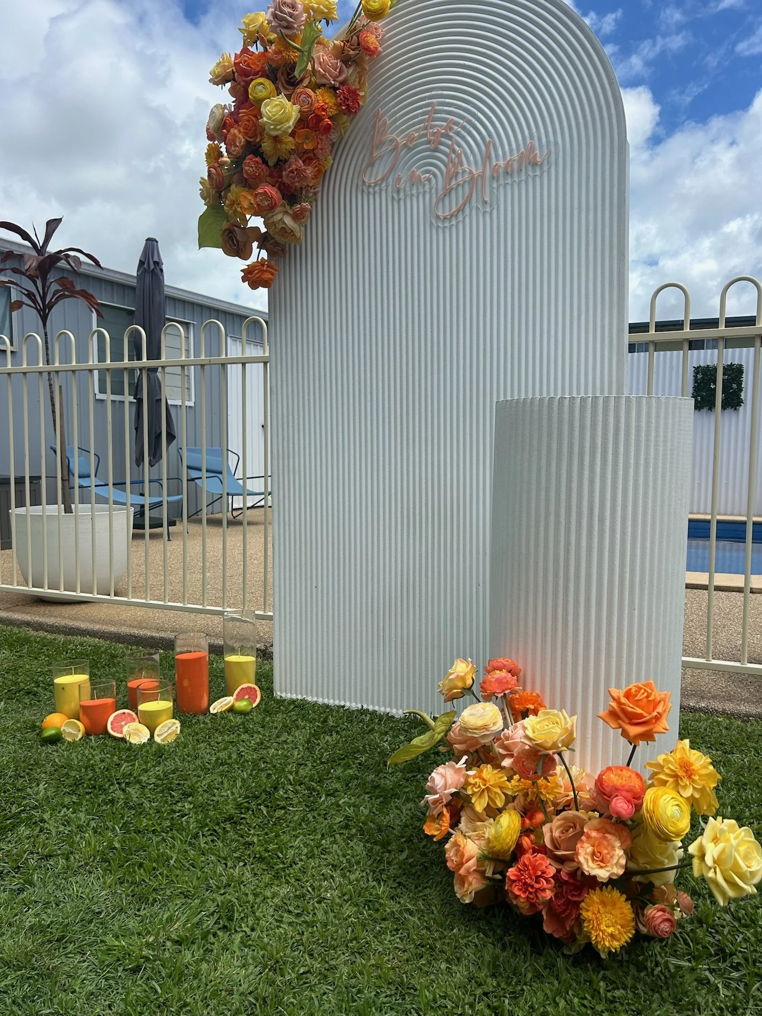 Decorative setup with a white textured panel, floral arrangements, candles, and a sign that says 'Bach' against a cloudy sky and outdoor setting.