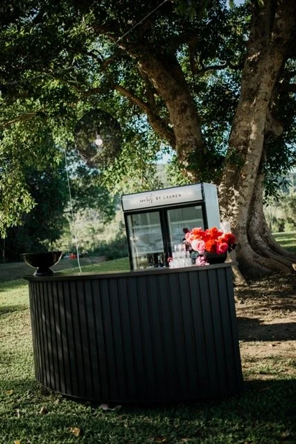 Outdoor black bar with a glass display case and a bouquet of pink roses, set under a large tree in a park.