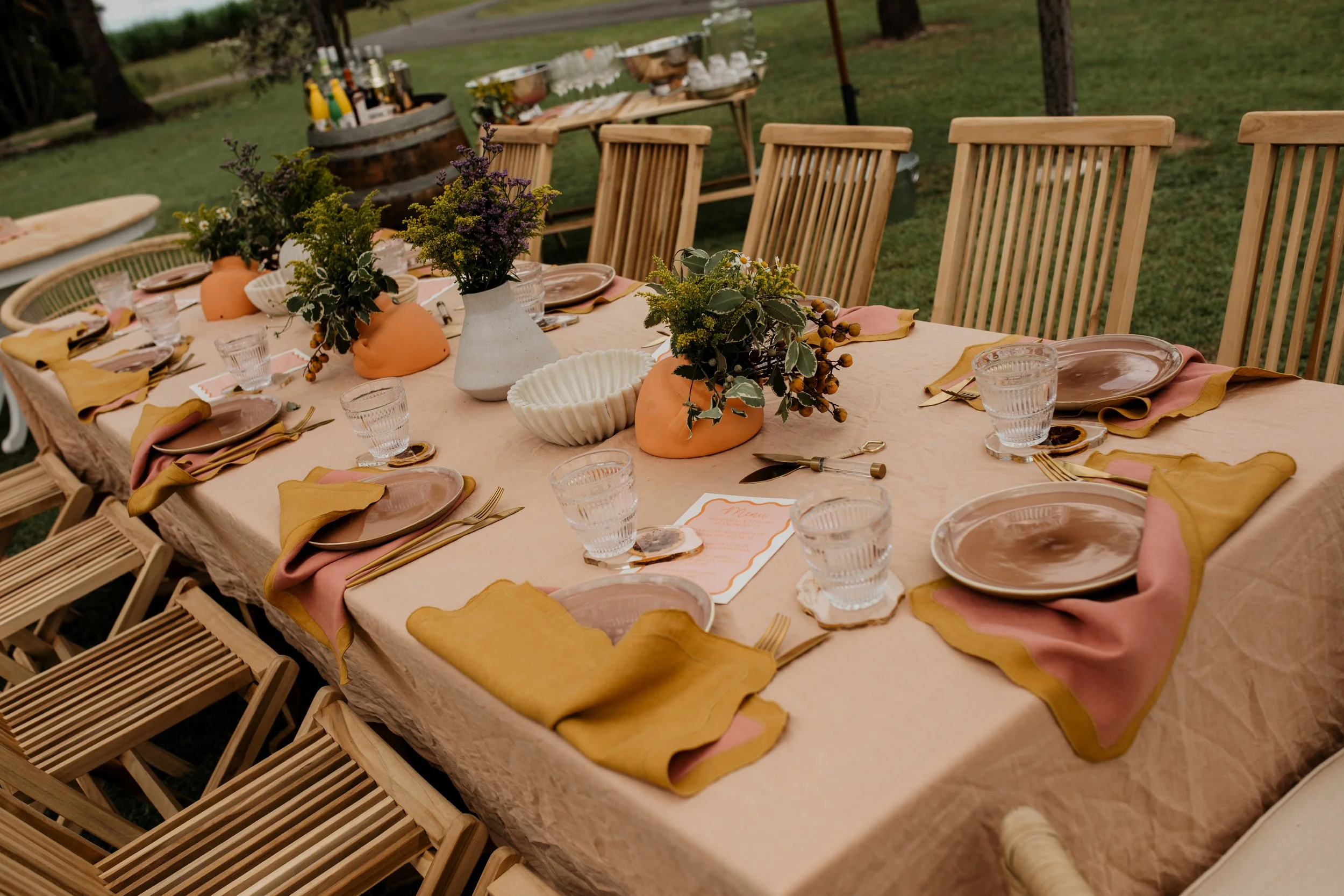 A beautifully set outdoor dining table with beige tablecloth, pink and mustard napkins, brown plates, and glassware. Centerpieces of flowers in white and orange vases are arranged along the table. In the background, there is food and drink setup on a