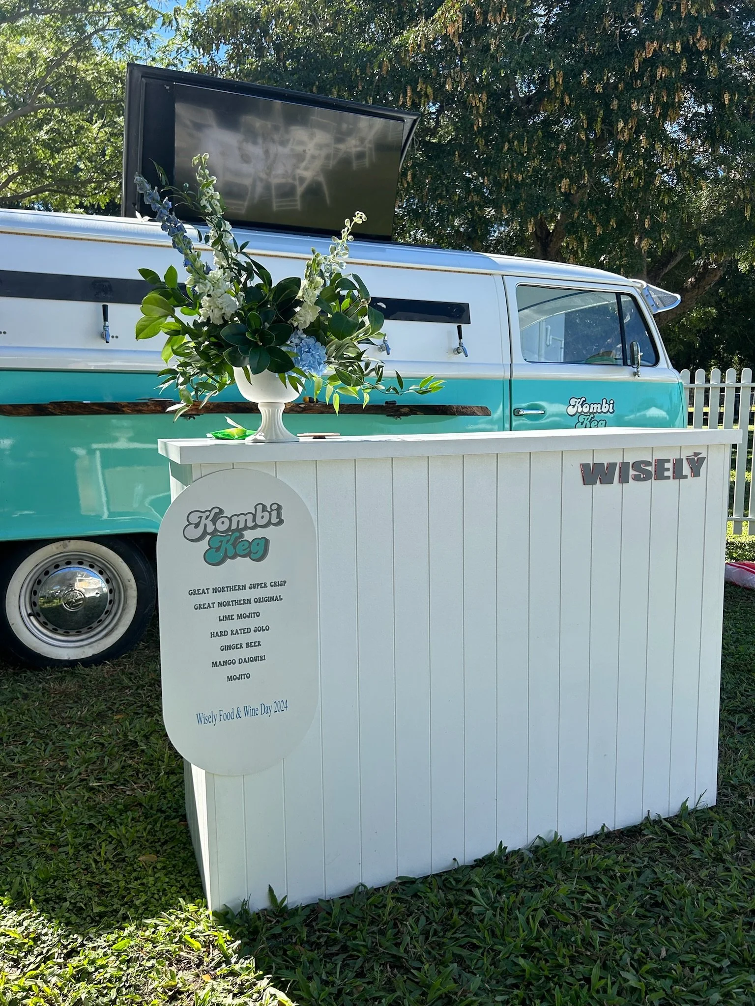 A teal and white vintage van with signage that reads 'Kombi Keg' and 'WISELY,' parked outdoors among green trees, with a white bar counter and a large floral arrangement in a white vase on top.