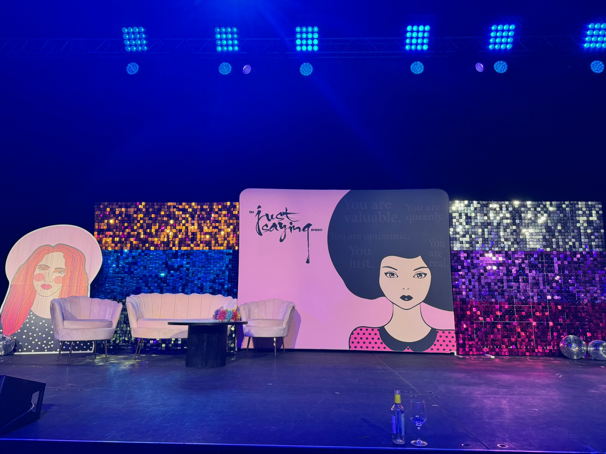 Stage setup with three pastel chairs and a table in front, decorated with a floral arrangement, with colorful sequin backdrops and illustrated banners including a woman with red hair and another with black hair, under blue stage lighting.