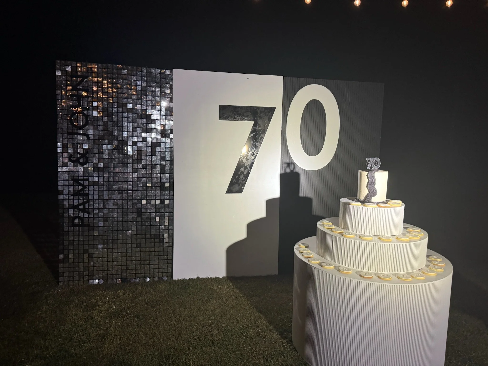 A celebration setup with a white tiered cake decorated with gold coins and a '70' topper, set against a backdrop with the number '70' and the words 'PASIÓN' and '72'.