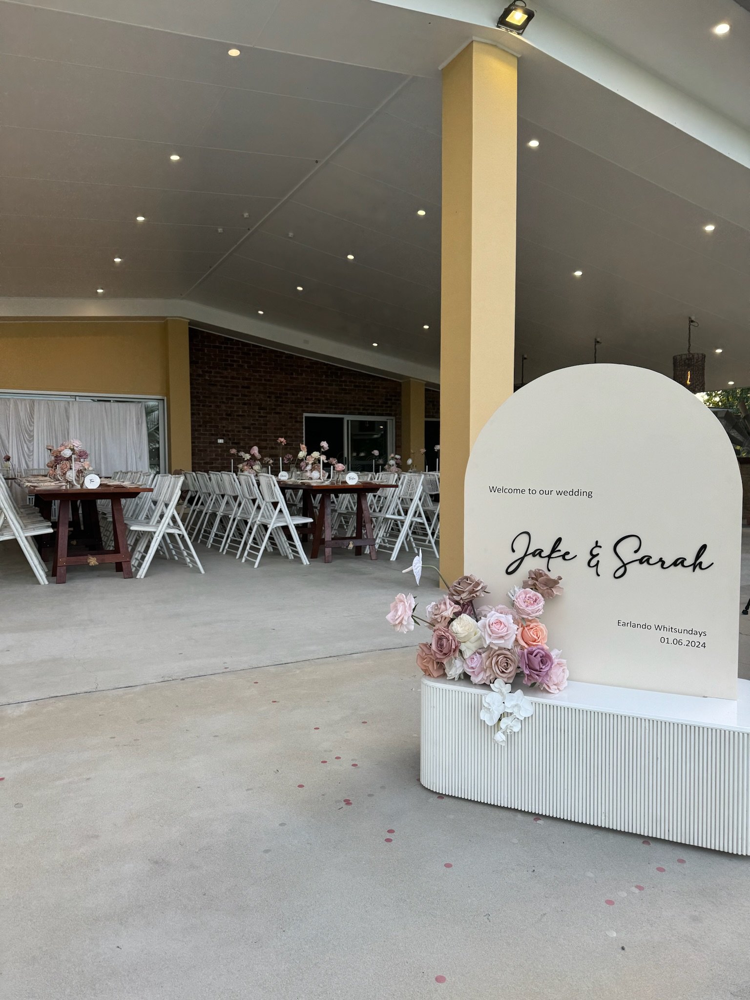 Wedding reception area with decorated tables and chairs, a large welcome sign with floral arrangements, and a covered outdoor space.