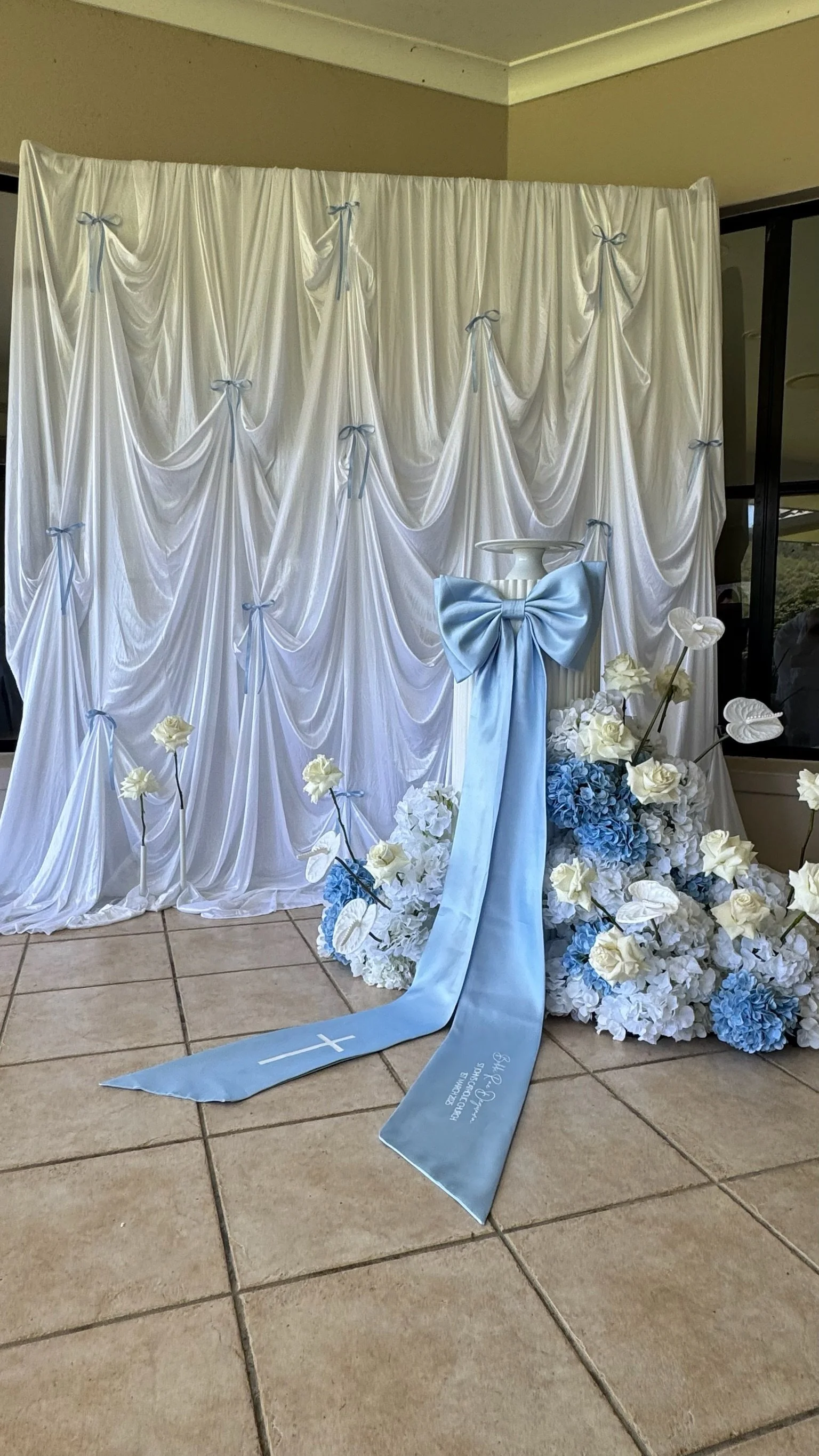 A white draped backdrop decorated with small blue ribbons, with floral arrangements of white and blue flowers and a white pillar with a large blue bow, and a blue sash with white embroidery and a cross, likely for a religious or wedding ceremony.