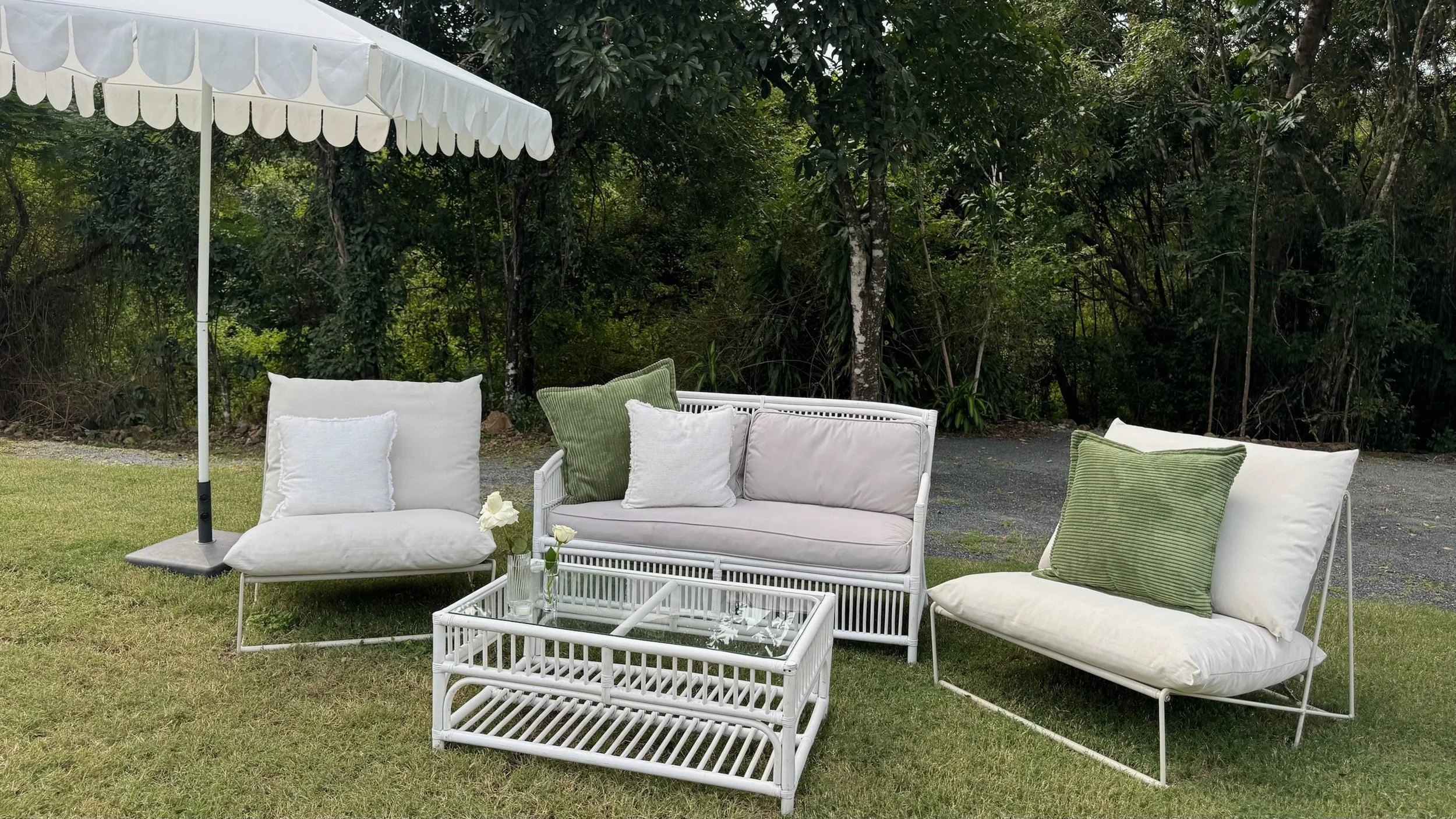 Outdoor patio furniture set including two cushioned chairs, a cushioned loveseat, and a glass-topped table with a vase of white flowers, all under a white patio umbrella on a grassy area with trees in the background.