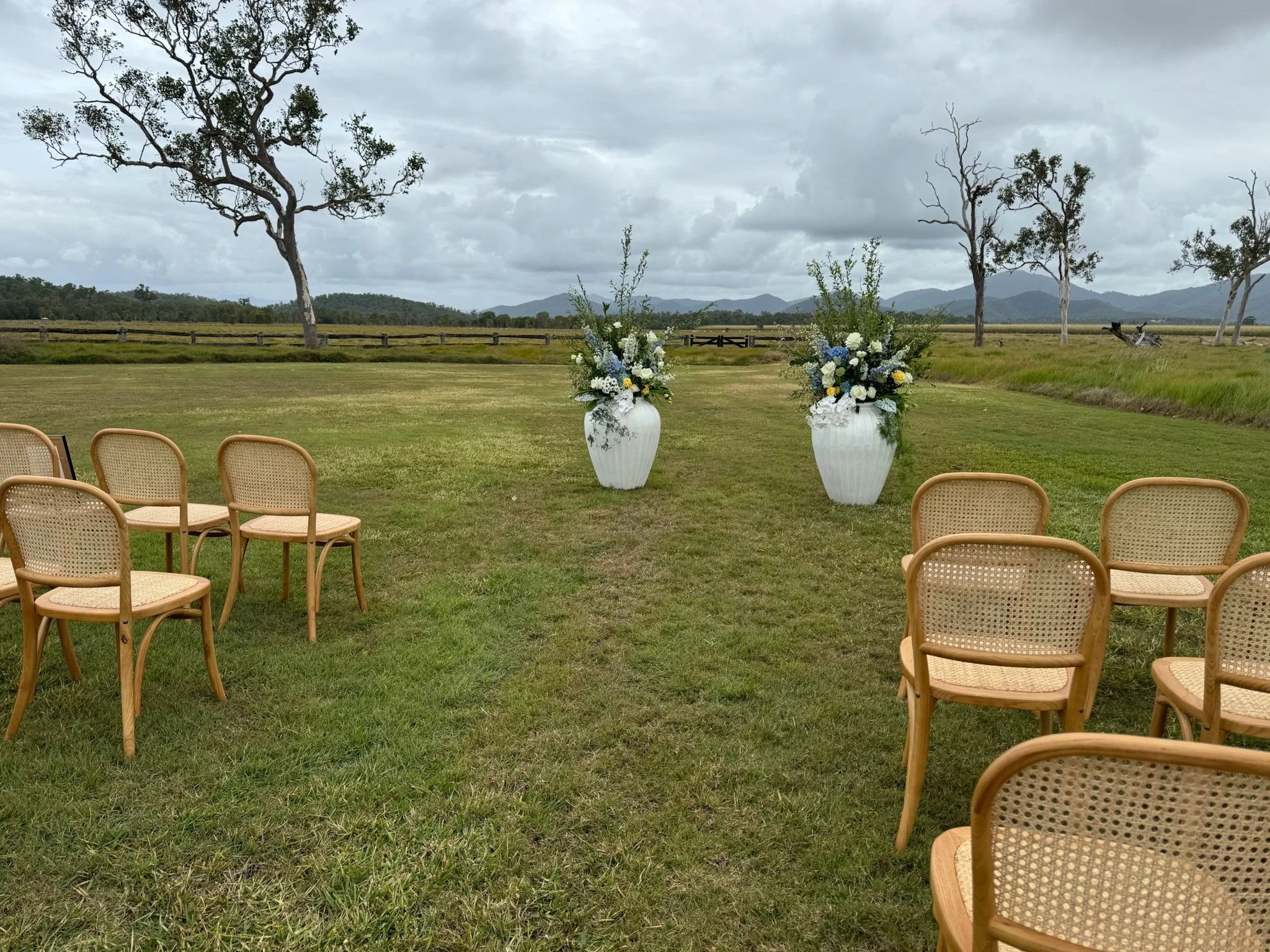 Outdoor wedding ceremony setup in a grassy field with two large white vases holding floral arrangements and light wooden chairs arranged on either side. Scattered trees and distant mountains under a cloudy sky.