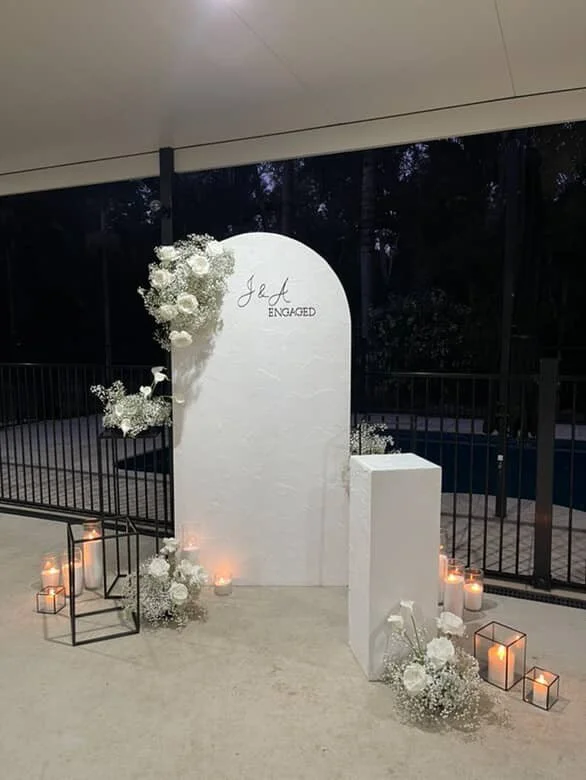 Wedding or engagement sign with floral decorations and candles, reading 'J & J ENGAGED', set up indoors with a black railing and large windows in the background.