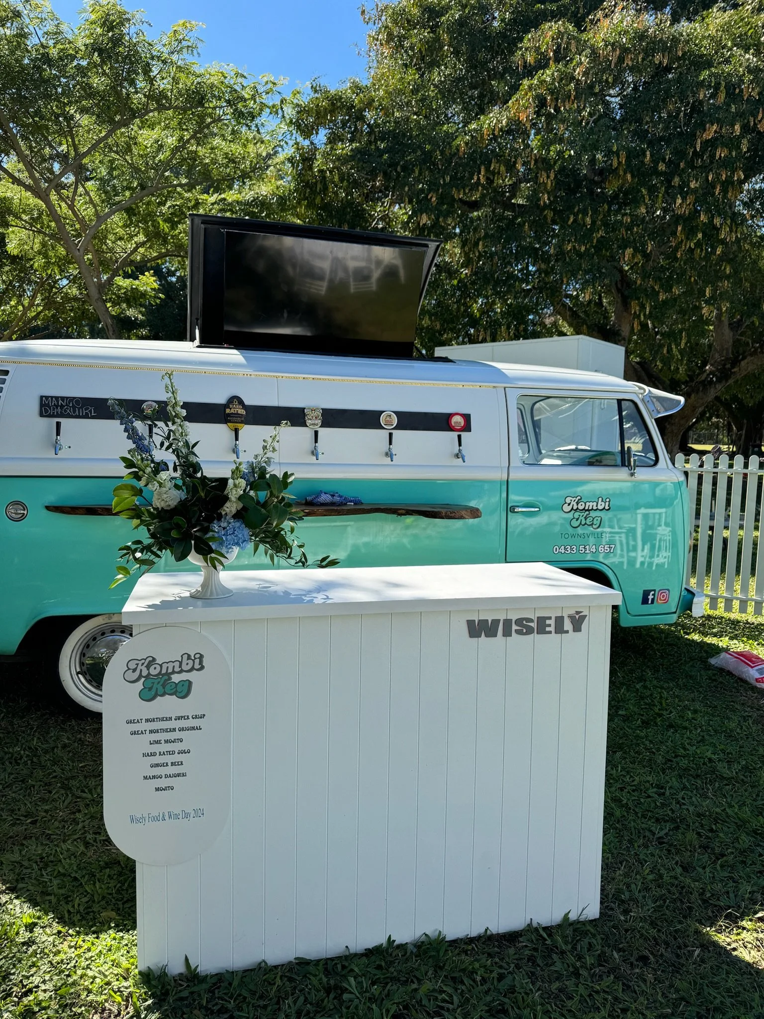 A vintage teal and white van with a large black screen on top and beer taps on the side, parked outdoors under trees. In front of the van, a white table and signboard display the menu for 'Kombi Keg,' featuring various beer options, and a floral arra
