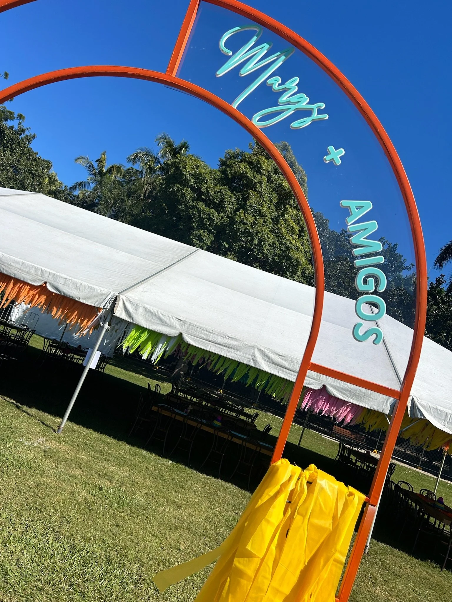 An outdoor event setup with a large white tent, decorated with colorful paper fringe, on a grass field under a clear blue sky. A decorative arch reads 'Margs + Antigos'.