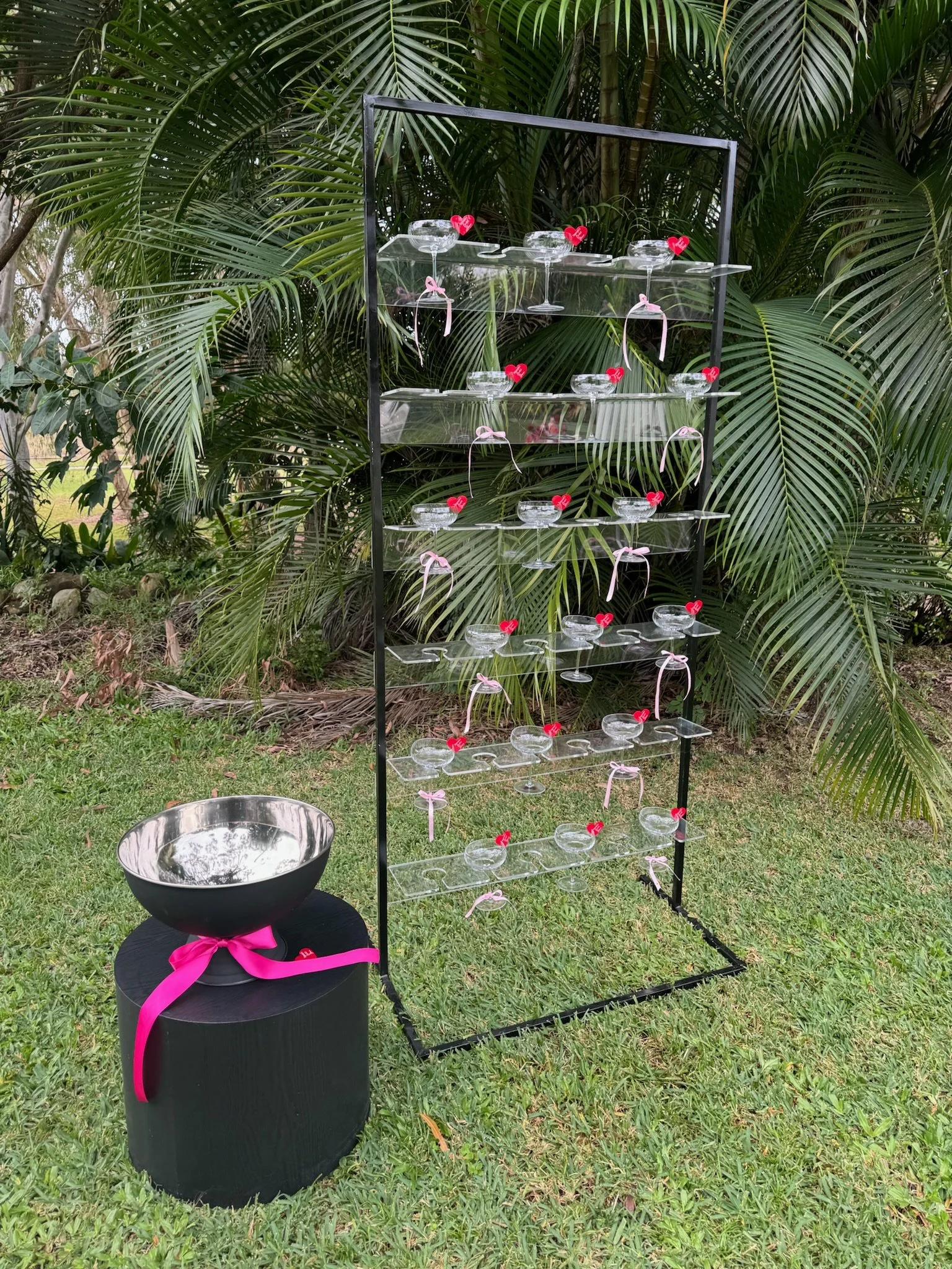 A display of empty glass cups on clear shelves, decorated with pink ribbons and small red heart-shaped toppers, set outdoors in front of lush green palm leaves, with a large metallic bowl with a pink ribbon on a black stand nearby.